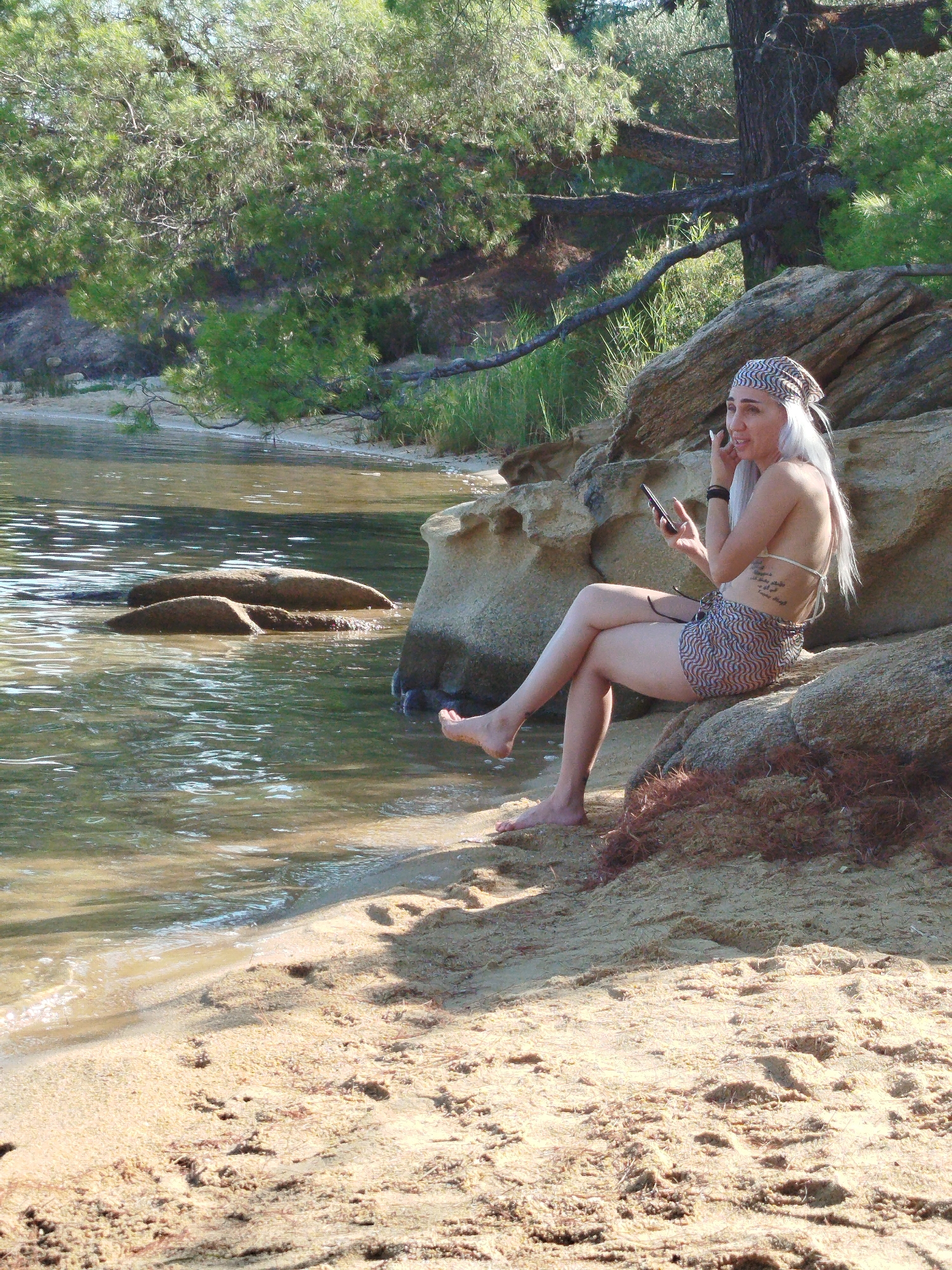 A woman with long, dyed platinum blonde hair and a patterned headscarf sits on rocks at the edge of a sandy beach. She is wearing a bikini top and patterned shorts. The woman is holding a mobile phone in her right hand and appears to be listening to something through earbuds. Her left hand is near her face, touching her ear. The water is calm and shallow, with some rocks visible just below the surface. The background shows dense green foliage and a large tree trunk. The location is likely a beach in Sithonia Municipal Unit, Greece.