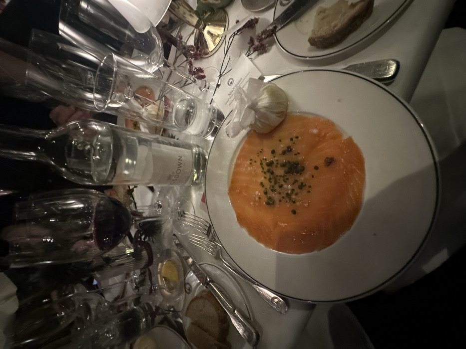 A table setting features a plate of salmon with capers and a lemon pouch. Beside it sits a bread plate with a slice of bread. A wine bottle labeled "NMODOWN" lies amidst multiple wine glasses. Silverware, including a fork and knife, are placed near the plates. The scene suggests an elegant dining experience.