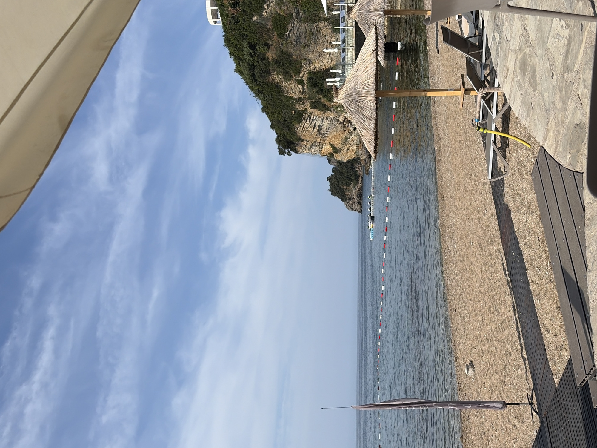 The image shows a beach scene in Budva, Montenegro. In the foreground, there are several beach chairs and straw umbrellas, suggesting a resort or public beach area. A yellow hose is visible on the sand. Wooden walkways are laid out on the sand. The sea is calm, with a safety line marked by red and white buoys. In the background, a rocky, tree-covered cliff rises from the sea, with some white structures visible at the top, likely part of the city's infrastructure. The sky is blue with scattered white clouds.