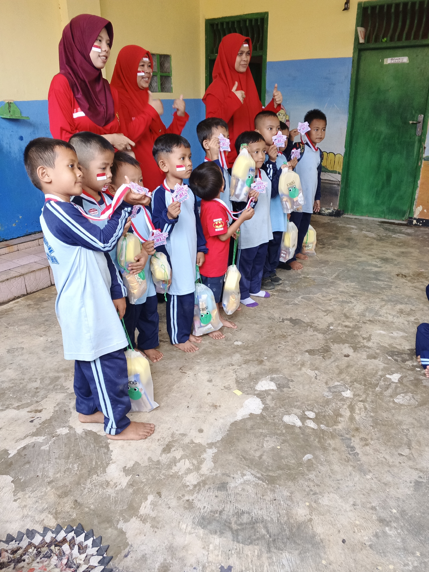 In Warungkiara, Indonesia, a group of young children, likely students, stand in a line outdoors, holding small bags. They are adorned with face paint resembling the Indonesian flag, indicating a patriotic event or celebration. Two adults, presumably teachers or guardians, stand behind the children, also wearing headscarves with Indonesian flag motifs. The children are dressed in uniforms, and some are holding medals. The ground appears to be a paved courtyard or schoolyard, with a building visible in the background. The overall scene suggests a school event, possibly a competition or national day celebration, taking place in Warungkiara.