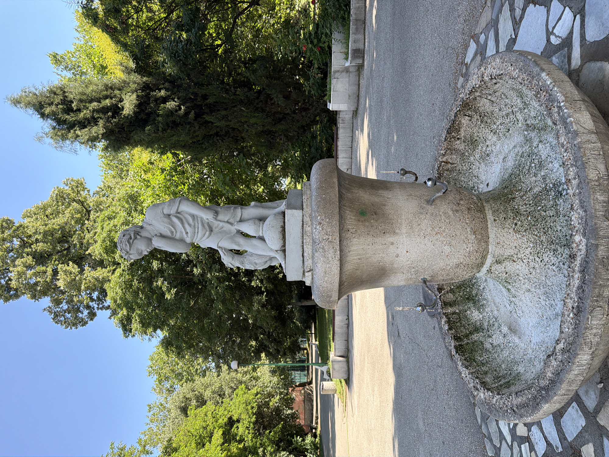 The image shows a weathered stone fountain in a park. A statue of a young boy holding a lamb stands atop the fountain, which has a decorative urn-shaped base and a basin filled with murky water. The fountain is surrounded by a pathway made of light-colored flagstones and gravel. In the background, lush green trees and a tall, slender evergreen tree create a natural setting. A lamppost and benches are visible in the distance, suggesting a public park. The sky is clear and blue. This fountain is located in Tsar Simeon's Garden in Pazardzhik, Bulgaria.
