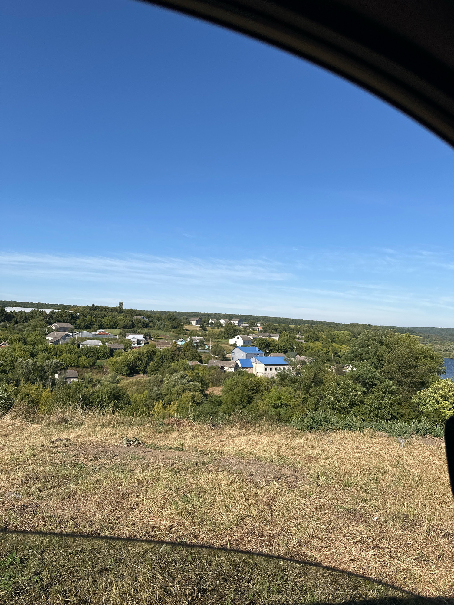 This is a landscape photograph taken from an elevated position, looking out over a rural village and surrounding greenery. The sky is a clear, bright blue with a few wispy white clouds. In the foreground, a dry, grassy slope dominates the lower part of the frame, with some scattered debris.
The middle ground features a village nestled amongst dense trees. The houses are varied in style and color, with some having visible roofs. Notably, several houses in the village have bright blue roofs. The village appears to be spread out, with houses interspersed with gardens and trees.
The background is characterized by a vast expanse of lush green trees, suggesting a heavily wooded area. A body of water, possibly a river or lake, is visible on the far right of the image, reflecting the blue sky. The overall impression is one of a peaceful, rural setting under a clear summer sky.
