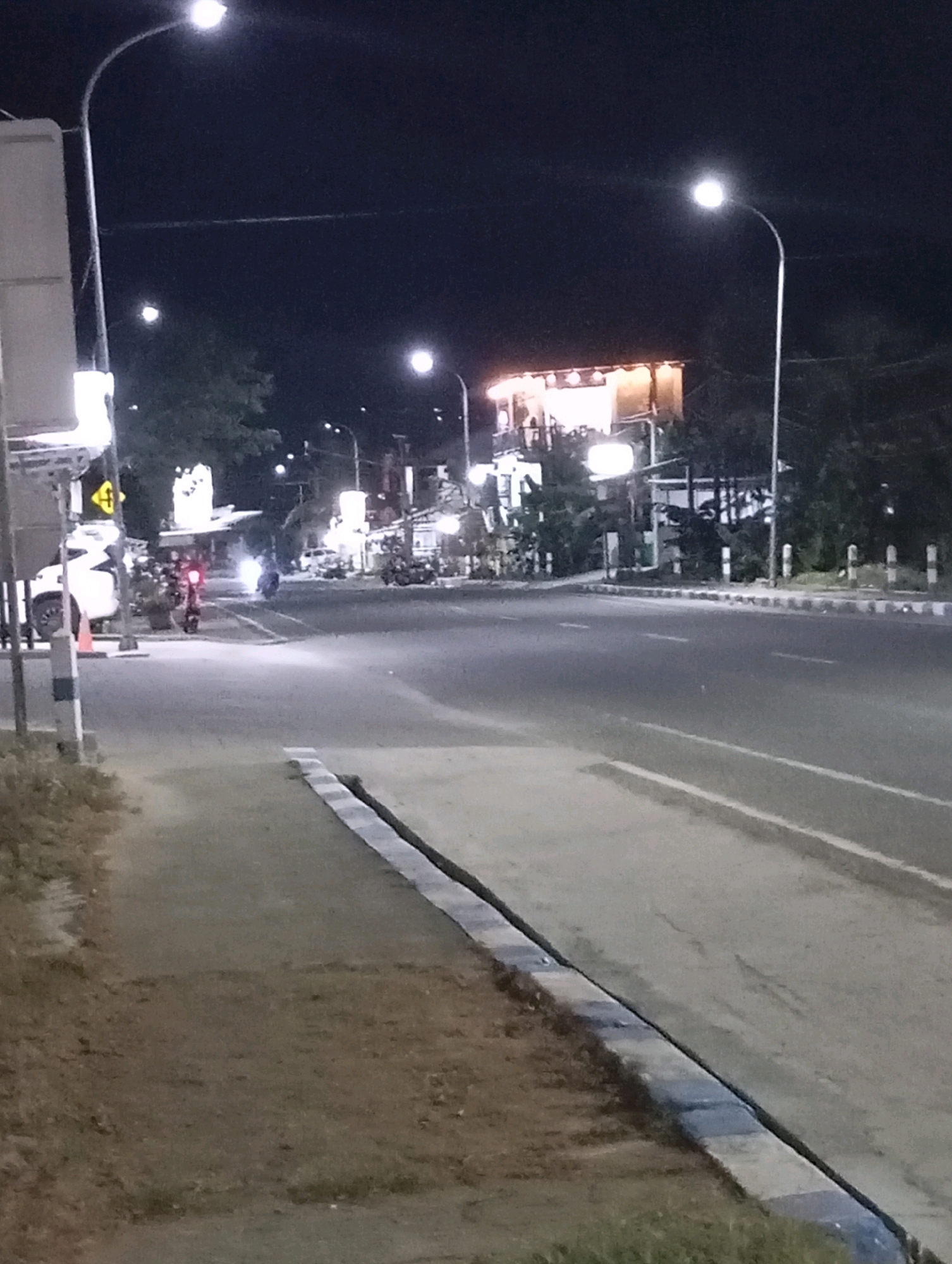 The image captures a nighttime street scene in Pacitan, Indonesia. A paved road occupies the majority of the frame, with a sidewalk running along the left side. Streetlights illuminate the scene, casting a glow on the road and surrounding trees. A building with illuminated windows is visible in the background. On the road, several motorcycles are present, some of which appear to be moving, with headlights and taillights visible. A white vehicle is parked on the left side of the road near the sidewalk. The overall atmosphere is typical of a street at night, with artificial lighting from streetlights and buildings.