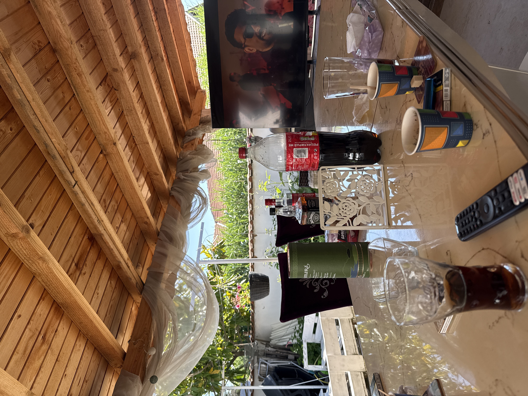The image depicts a scene likely taken outdoors in Elhovo, Bulgaria. The foreground features a table covered in various items including drinks (Coca-Cola, Whiskey), glasses, a television screen showing a person, a remote control, and other miscellaneous objects. The table sits beneath a wooden structure suggesting a patio or gazebo. Through the gaps in the wooden structure, a glimpse of the neighborhood with houses and greenery is visible. No people are directly visible in the image.