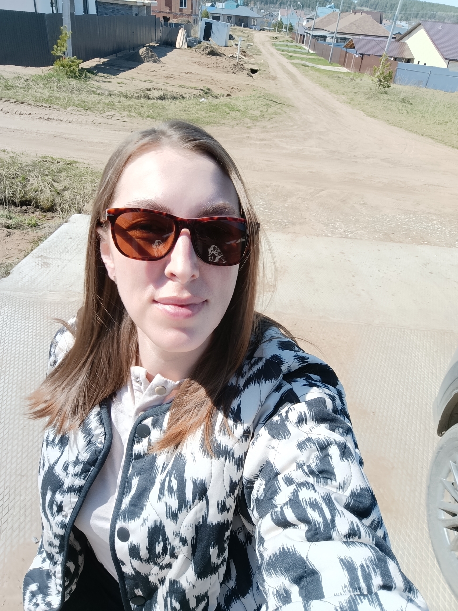 A fair-skinned woman with long brown hair and brown sunglasses is taking a selfie outside in Chaikovsky, Russia. She is wearing a white shirt with buttons and a black and white patterned jacket. In the background, there is a dirt road leading to a residential area with houses of various sizes and colors. Construction appears to be ongoing in the area. The weather appears to be sunny. A car wheel is partially visible in the lower right corner of the image.