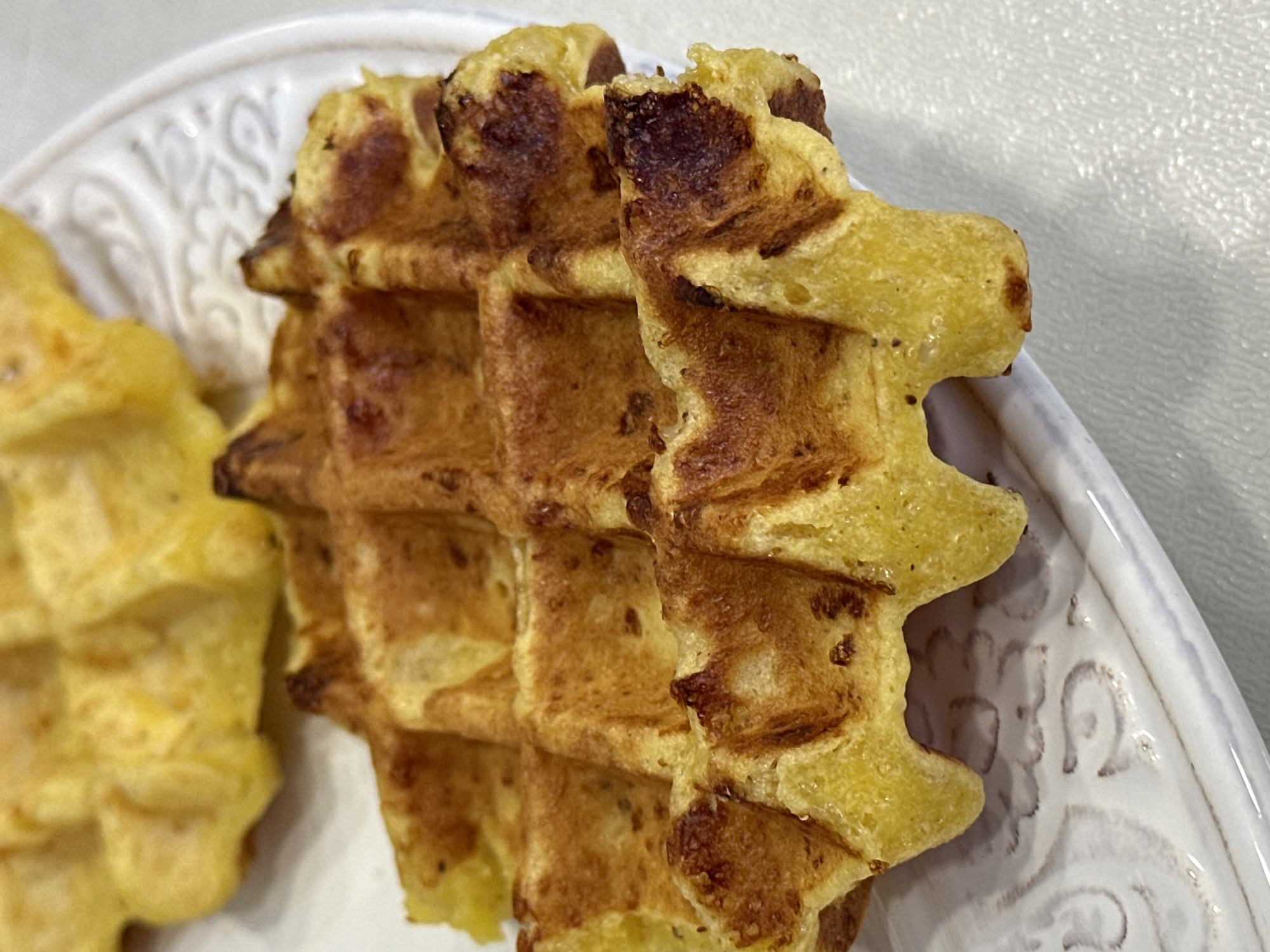 The image shows a close-up of two golden-brown waffles served on a white, embossed plate. The waffles have a distinct grid pattern, and some edges appear slightly charred, suggesting they are well-cooked. The waffle in the foreground is larger and sharper in focus than the one behind it, which is partially visible and slightly out of focus. The plate is set on a light-colored, textured surface, possibly a tablecloth or countertop. The overall impression is one of a simple, homemade breakfast or snack. The location is not specified in the image or context.