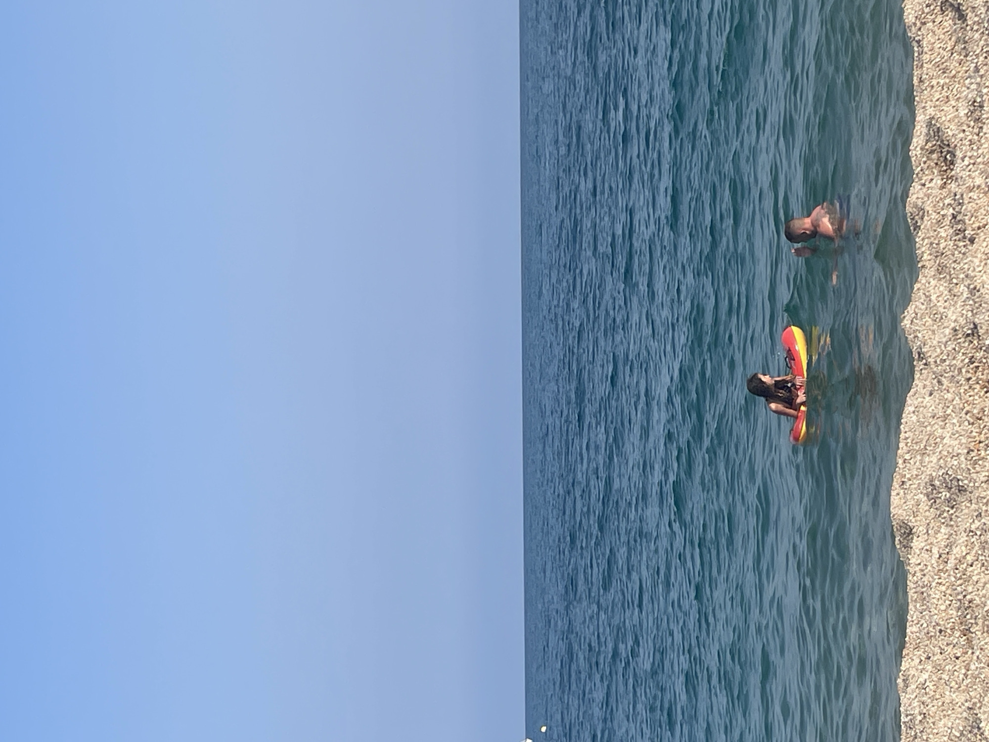 Two individuals are in the ocean at Arenys de Mar, Spain. In the foreground, a person is partially submerged in the water, with their upper body and head visible. Further back, a child is seated on a red and yellow inflatable ring, also in the water. The ocean is calm with small waves, and the sky above is clear and blue. The shoreline is composed of small pebbles.