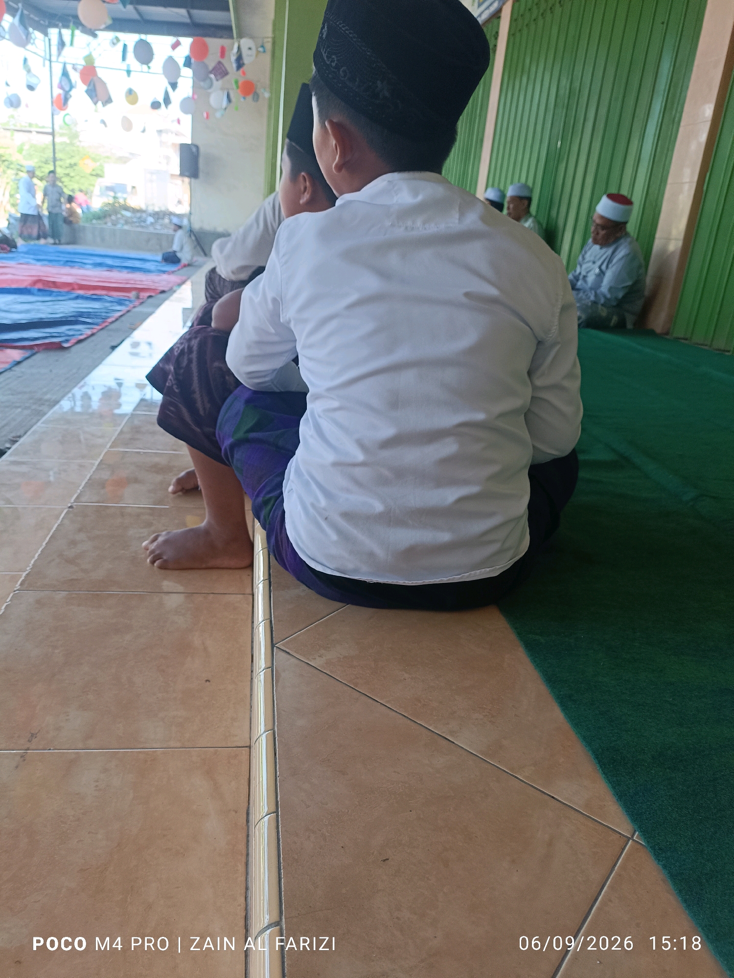 The image shows two young boys in traditional Islamic clothing, including kufi caps and long white shirts, sitting on a tiled floor. One boy is facing away from the camera, while the other is partially visible from the side. In the background, several other individuals are seated on a green carpet, some wearing kufi caps and peci hats. The environment appears to be an outdoor or semi-outdoor space, with a green corrugated metal wall and a covered roof structure visible. Decorations, possibly balloons and flags, hang from the ceiling in the distance, suggesting a celebratory event. The scene takes place in Cendagah, Indonesia, as indicated by the timestamp overlay.