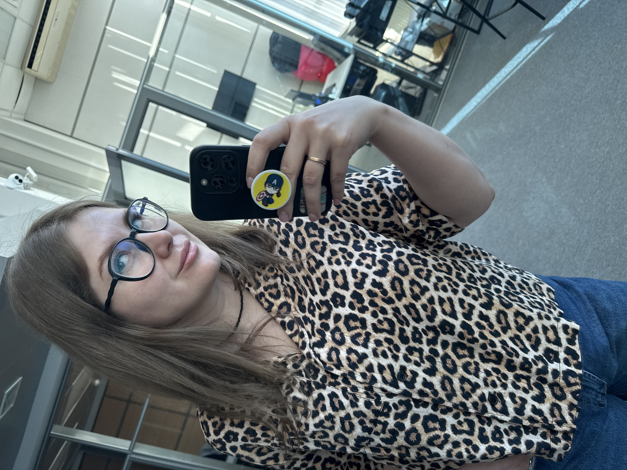 A person with light brown hair and glasses is taking a selfie. The person is wearing a leopard print shirt and blue jeans. The phone they are using is black with a circular pop socket featuring a cartoon character resembling Captain America. The location appears to be an indoor space with large windows, possibly an office or waiting area, with chairs and other furniture visible in the background. Based on the information available, determining the precise location in the city of Perm, Russia, is not possible.