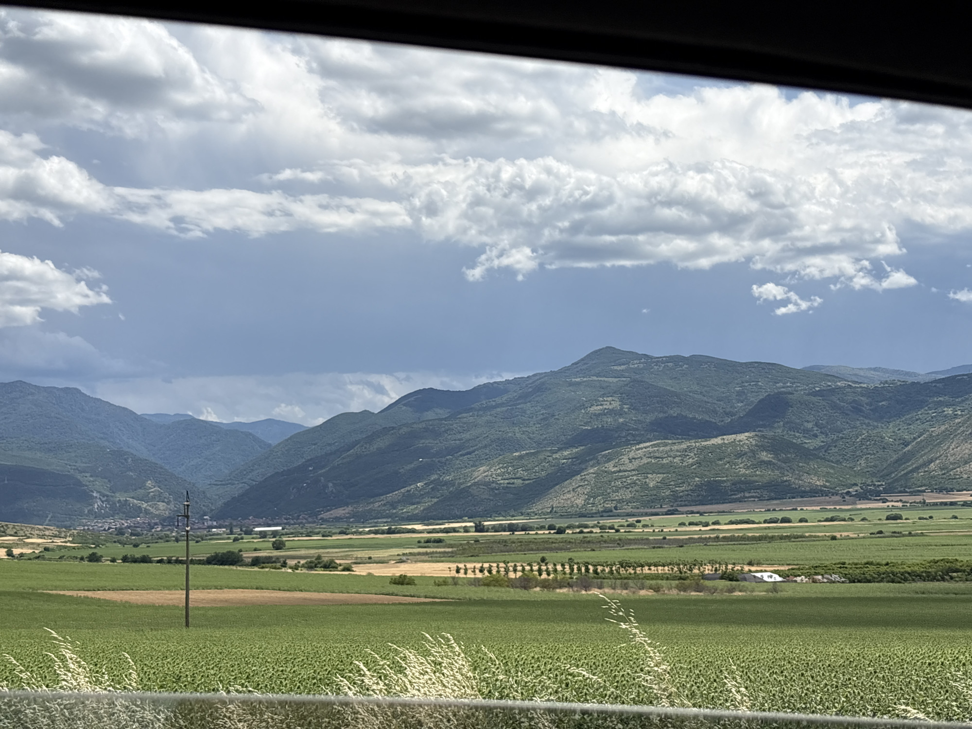 The image presents a wide-angle view of a rural landscape, likely in or near Isperihovo, Bulgaria, captured from a moving vehicle. In the foreground, a vibrant green field of crops, possibly sunflowers or a similar low-lying plant, occupies the lower portion of the frame, with wispy dry grass blades visible along the bottom edge, suggesting a barrier or edge.
Midground features include several fields with varying shades of green and golden-brown, indicating different crops or stages of growth. A single, weathered wooden utility pole stands prominently in the middle of one of these fields. Further back, a line of trees creates a natural boundary, behind which lies a cluster of buildings that appears to be a small village or settlement nestled in the valley.
The background is dominated by rolling, verdant mountains, their slopes covered in dense vegetation. The sky above is dramatic, with a mix of bright white cumulus clouds and darker, heavy storm clouds, hinting at impending or recently passed rain. The lighting is diffused due to the cloud cover, casting subtle shadows across the landscape. The overall impression is one of a vast, agricultural expanse under a dynamic sky, with the mountains providing a scenic backdrop.