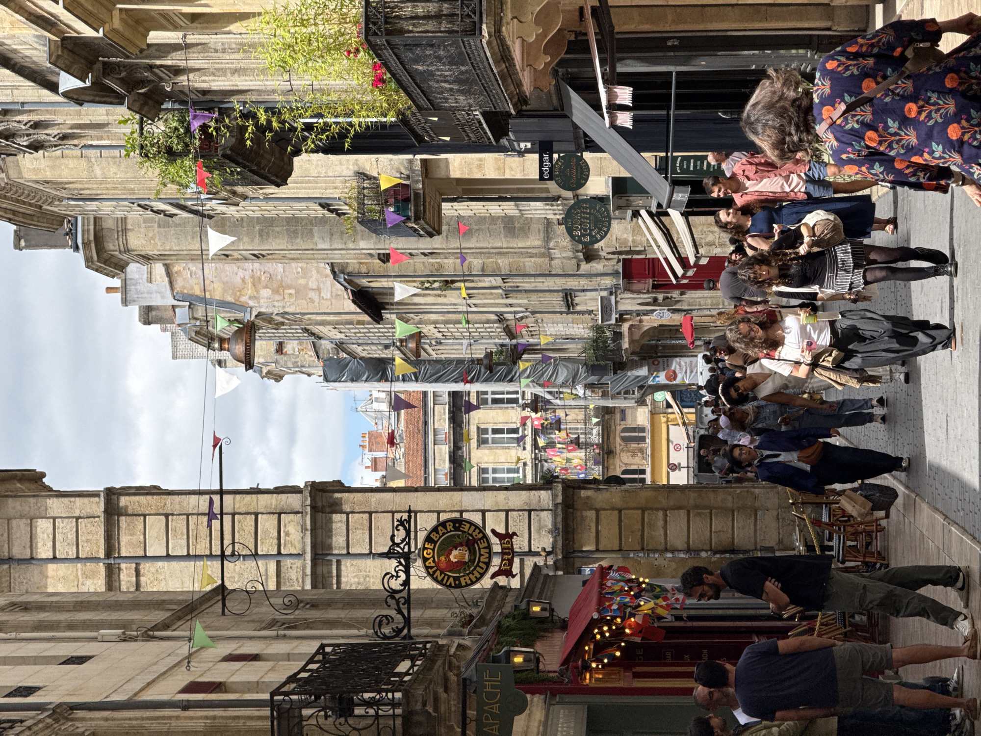 The image depicts a bustling street scene in Bordeaux, France, likely a pedestrianized area with narrow buildings lining both sides. The architecture is characterized by light-colored stone facades, with several balconies adorned with greenery and flags. Decorative bunting and strings of small flags are strung across the street, adding a festive atmosphere. On the left side of the street, a sign for "APACHE" is visible above an entrance with an awning decorated with numerous small international flags. Further down, a round sign reads "VINTAGE BAR RHUMERIE PUB". On the right side of the street, signs for "edgar." and "LES BOOKS & COFFEE BORDEAUX" are present. People are walking in both directions along the cobblestone street, engaged in various activities such as shopping, conversing, and simply passing through. The lighting suggests it is daytime, with some areas in shadow cast by the buildings. The overall impression is of a vibrant urban environment, possibly a commercial or historic district.