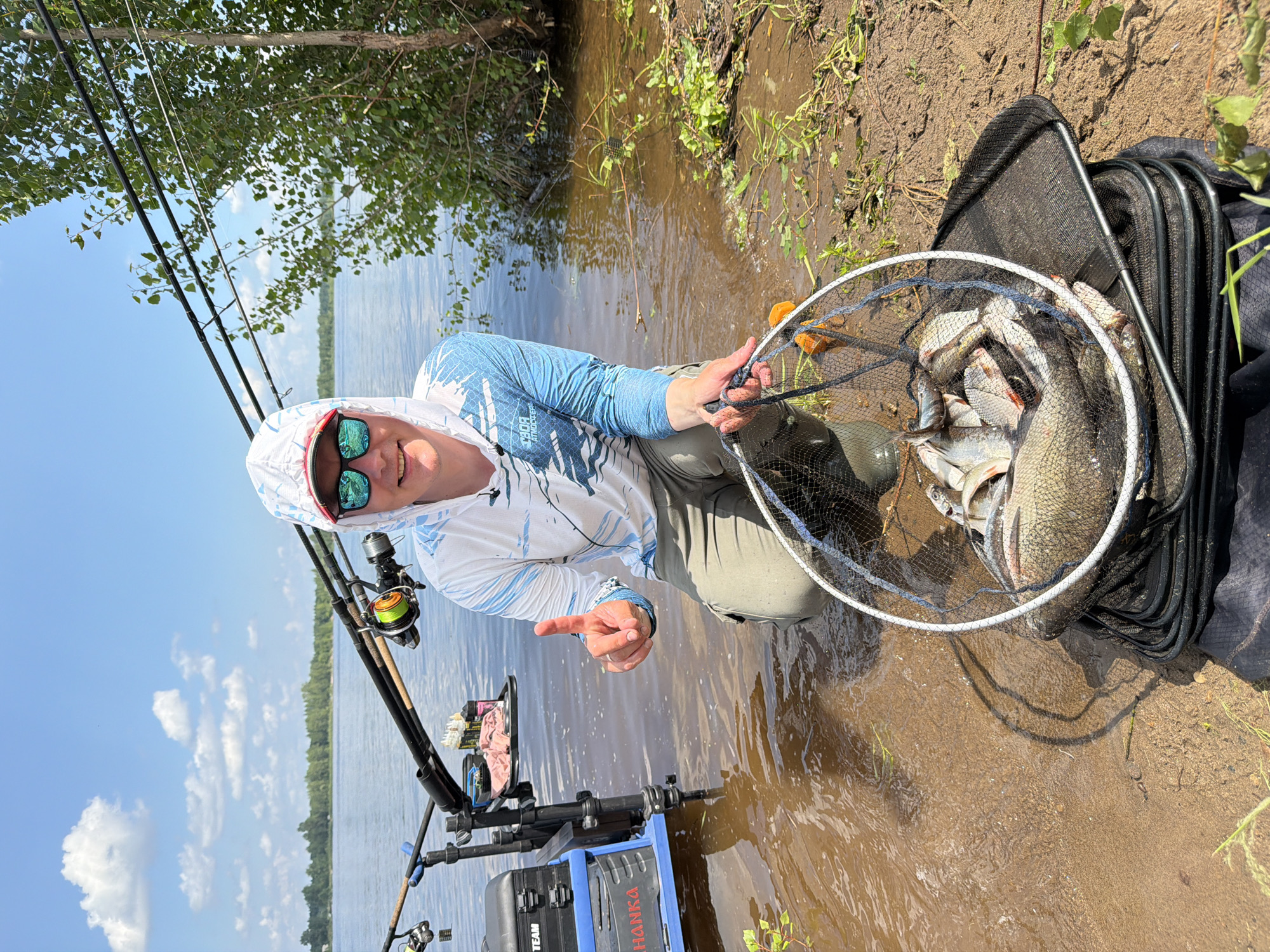 A Caucasian male is crouched by the water's edge, holding a fishing net filled with fish. He is wearing a white and blue long-sleeved shirt with a hood, sunglasses, and tan pants. He is smiling and giving a thumbs-up gesture. In the background, there are fishing rods set up on a stand, a blue fishing box, and a body of water. The environment appears to be a natural setting with trees and vegetation. The overall impression is that of a successful fishing trip. The city of Tarasovo, Russia is mentioned in the prompt as the location.