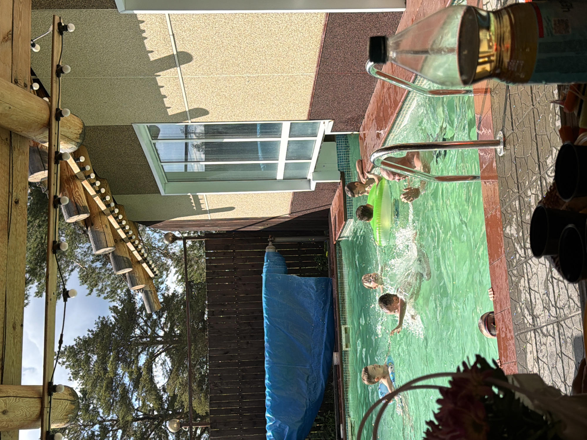 A group of children and an adult are enjoying a sunny day at an outdoor swimming pool. The pool is rectangular with a tiled edge and is filled with clear, green water. Several children are visible in the water, some with inflatable rings. One child is splashing, while another is partially submerged, leaning on the pool edge. An adult male is in the pool with his hands raised. In the background, a wooden fence encloses the pool area, and a blue tarp is partially covering a rolled-up pool cover. The scene appears to be a residential backyard, likely in a suburban or rural setting, potentially in the city of Chaikovsky, Russia, given the context. The architecture of the building adjacent to the pool suggests a rustic design with exposed wooden beams and textured walls. Overhead, string lights are hung from the eaves of the building, indicating a potential for evening use. The overall atmosphere is one of leisure and recreation.