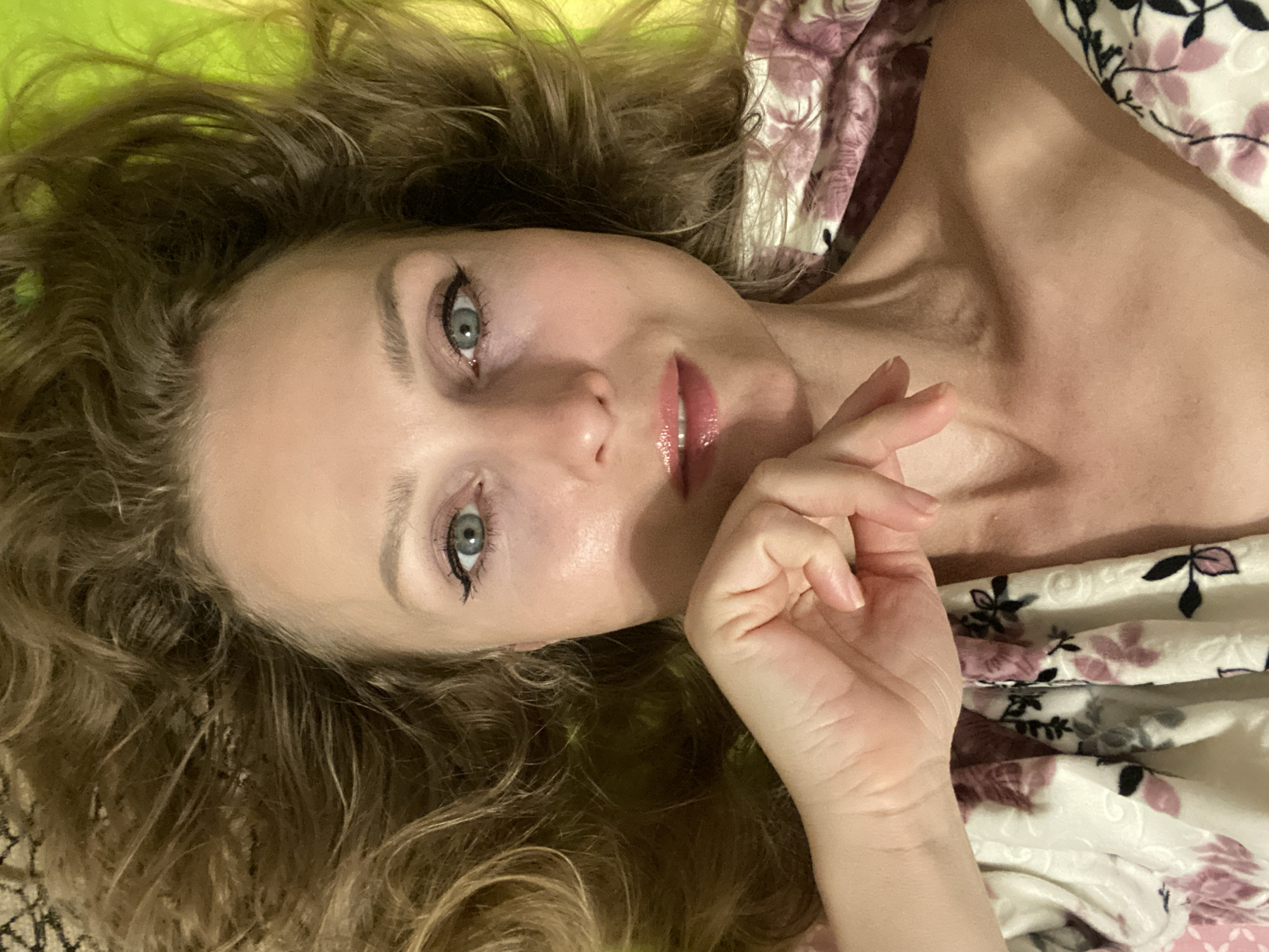 This is a close-up, overhead, selfie-style image of a woman lying down. She has blonde, wavy hair fanned out around her head. Her eyes are open and looking directly at the camera. She is wearing eyeliner and mascara. Her lips are glossed. Her right hand is resting on her chin. She is wearing a garment with a floral pattern. The background appears to be a textured surface with a bright green area visible. The overall lighting suggests an indoor setting. The image does not contain any information about Lida, Belarus.
