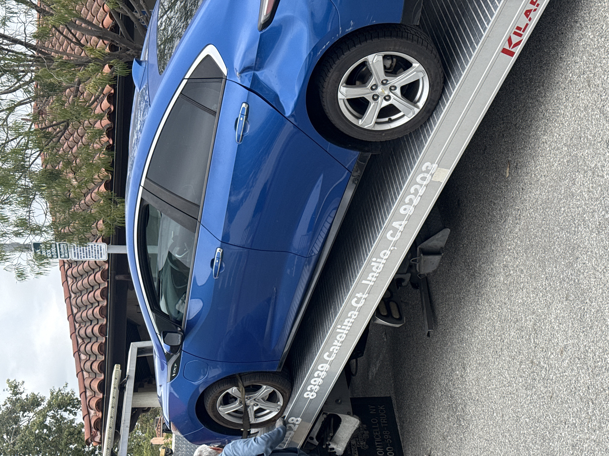 A damaged blue Chevrolet Volt is being towed on a flatbed tow truck. The car has a noticeable dent near the passenger side rear door. The tow truck displays text that includes "83939 Carolina Ct Indio, CA 92203", indicating a location in Indio, California, rather than Los Angeles. The brand name "KILAR" is also printed on the truck. A person wearing a light blue shirt is partially visible near the front of the truck, possibly assisting with the towing operation. In the background, there is a building with a red tile roof and a sign.
