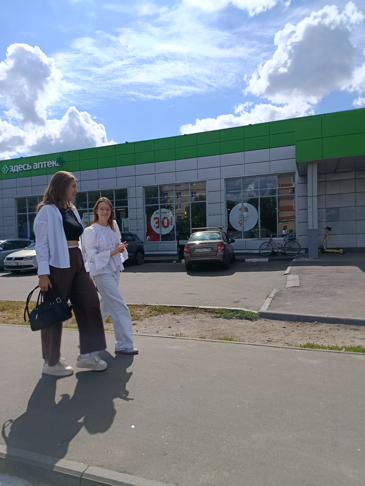 Two young women are walking on a sidewalk in front of a modern building with a green and silver facade. The building appears to be a pharmacy, as indicated by the text "ЗДЕСЬ АПТЕКА" (Here Pharmacy) in Russian on the upper left of the building. The woman on the left is wearing a white button-down shirt over a black crop top and striped brown pants, carrying a black handbag. The woman on the right is wearing a white ruffled shirt and white pants, and she is smiling and looking at the camera. In the background, there are cars parked in front of the building, including a brown Lada and a silver sedan. A bicycle is leaning against the building, and a yellow scooter is parked nearby. The sky is blue with scattered white clouds, suggesting a sunny day. The scene is set in a city environment, likely Orekhovo-Zuyevo, Russia, based on the context provided.