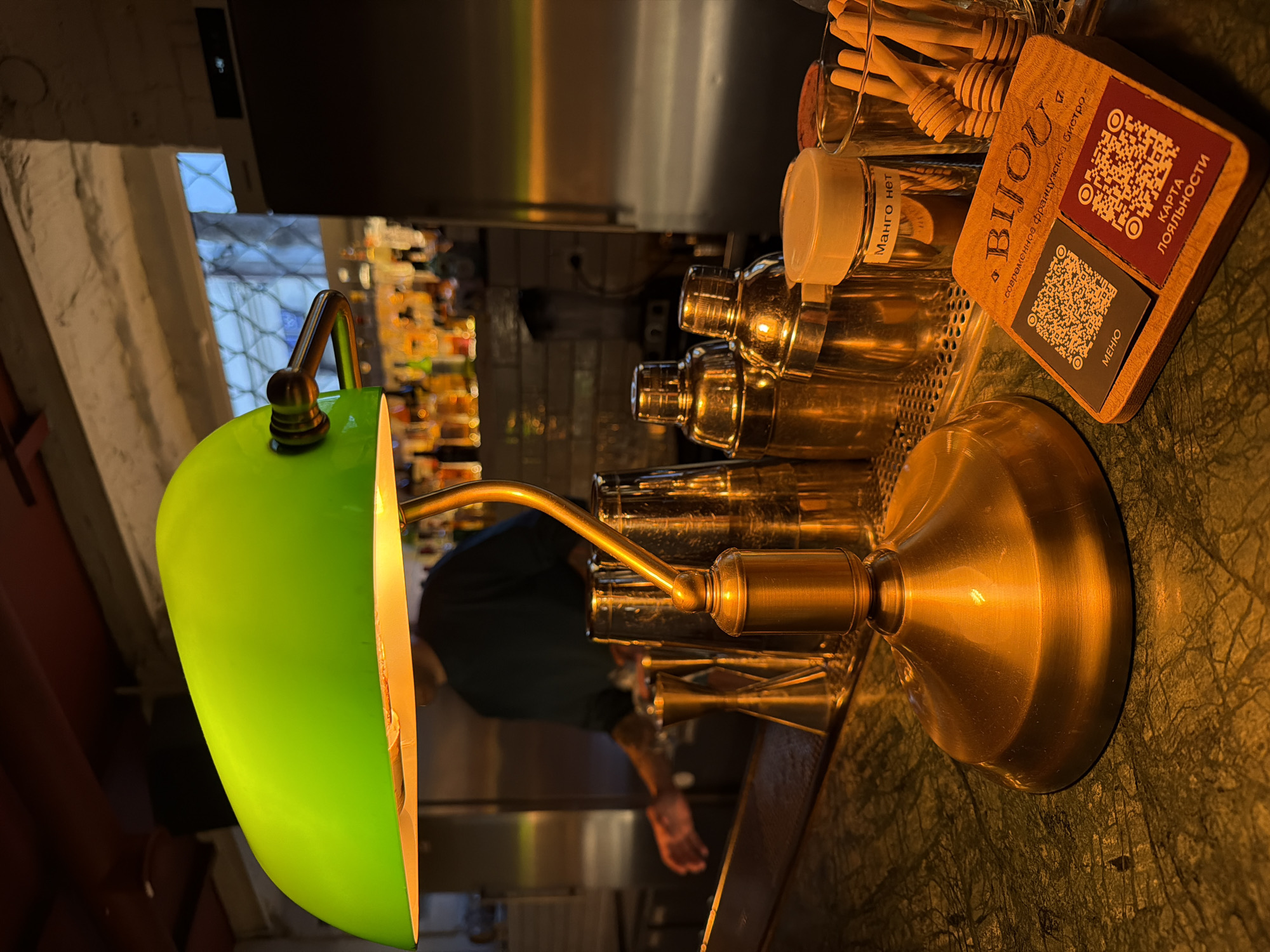 The image shows a bar countertop in an unidentified location. A bright green desk lamp with a gold base sits on a green marble countertop. Behind the lamp, there are several cocktail shakers, glass jars, and a wooden holder with honey dippers and a branded QR code card for "BIJOU" (a modern French bistro). In the background, shelves are stocked with bottles, suggesting it is a bar setup. A person wearing a dark shirt is faintly visible behind the cocktail shakers, possibly working at the bar. The lighting is warm and artificial, creating a cozy atmosphere.