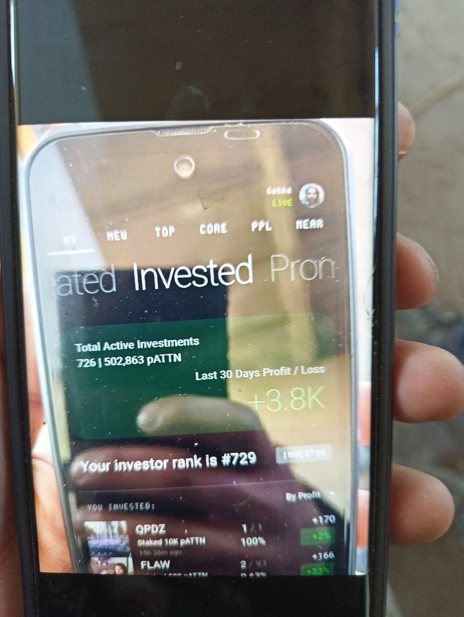 The image shows a close-up of a smartphone screen displaying an investment app. The app interface features metrics such as "Total Active Investments," indicated as 726 | 502,863 pATTN, and "Last 30 Days Profit/Loss" showing +3.8K. The user's investor rank is listed as #729. Below this, two investment options, "QPDZ" and "FLAW," are displayed, along with profit and stake percentages. The surrounding context includes a hand holding the smartphone. The location of the image is not discernible from the available information.