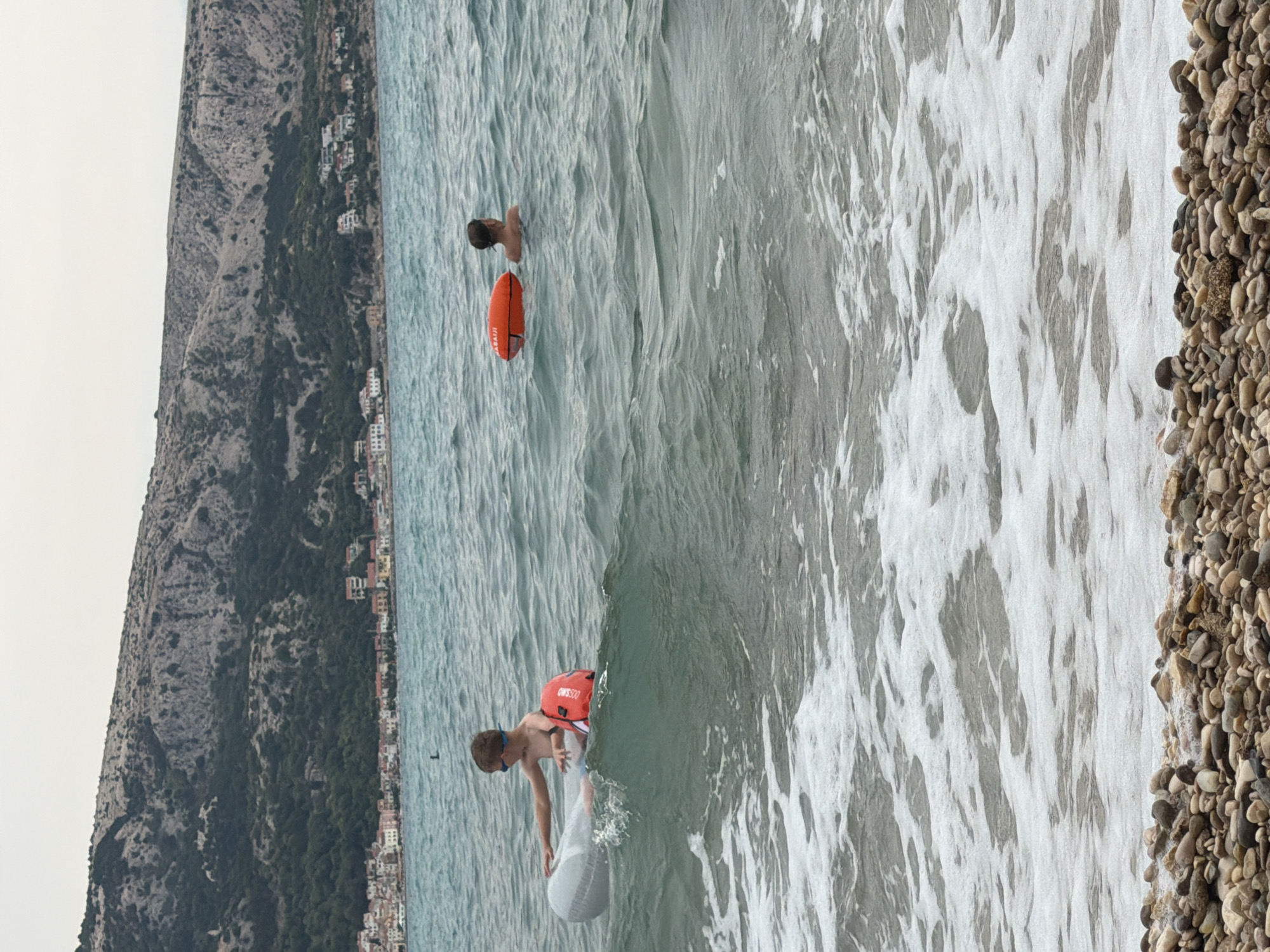 The image shows a boy in the sea wearing an orange life vest and blue goggles. He is holding onto a white float or raft. In the background, another person is visible in the water with an orange float. The scene is set on a pebble beach with the sea to the right of the frame and a mountainous coastline with buildings in the background to the left. The water is choppy with waves breaking and creating foam. The sky appears overcast, suggesting a cloudy day. The location is identified as Općina Baška, Croatia. The boy's body language suggests he is actively engaged in swimming or playing in the water. The text "ASSISTO" is visible on the orange life vest.