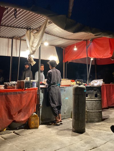 **Scene Description:**
An outdoor street food stall in Kandahar, Afghanistan. The stall is constructed with corrugated metal roofing and draped with white and red fabric. Three men are present: two stand behind the counter, appearing to be customers, while the third man, wearing a dark robe, stands in front of the counter, possibly a vendor. He is seen holding something in his hands. A large gas cylinder and what appears to be a metal barrel are present near the stall, likely used for cooking purposes. The scene is lit by hanging light bulbs from the roof of the stall. A yellow plastic container sits under the counter. The surface the stall rests on appears to be paved. The environment suggests it is nighttime or dusk.