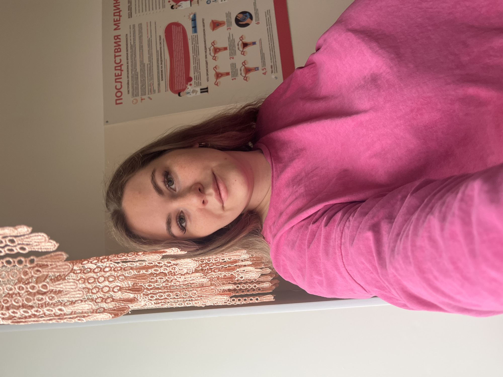 A young woman with light brown hair and a neutral expression looks directly at the camera in what appears to be an indoor setting. She is wearing a bright pink long-sleeved top. To her left, a decorative, intricately patterned lace-like textile in a light peach or beige color is visible.
Behind her, on a light-colored wall, hangs a medical or educational poster written in Russian. The prominent title at the top reads "ПОСЛЕДСТВИЯ МЕДИК" (Consequences of Medical), likely referring to medical abortion given the context of the subheadings. Sections on the poster include "НАСТРОЙКА ОРГАНОВ ВО ВРЕМЯ БЕРЕМЕННОСТИ" (Adjustment of organs during pregnancy) and "ВЛИЯНИЕ НА ЭНДОКРИННУЮ СИСТЕМУ" (Influence on the endocrine system). A significant section asks "ЧТО ПРОИСХОДИТ ПРИ МЕДИКАМЕНТОЗНОМ АБОРТЕ" (What happens during medical abortion). The poster features numbered diagrams (1 through 6) illustrating female reproductive anatomy, specifically the uterus and ovaries, depicting changes or processes relevant to the discussed topics.
The setting, with its medical poster, suggests a clinic, doctor's office, or a health education environment in Chaikovsky, Russia. The lighting is artificial and generally even, indicating an indoor location.