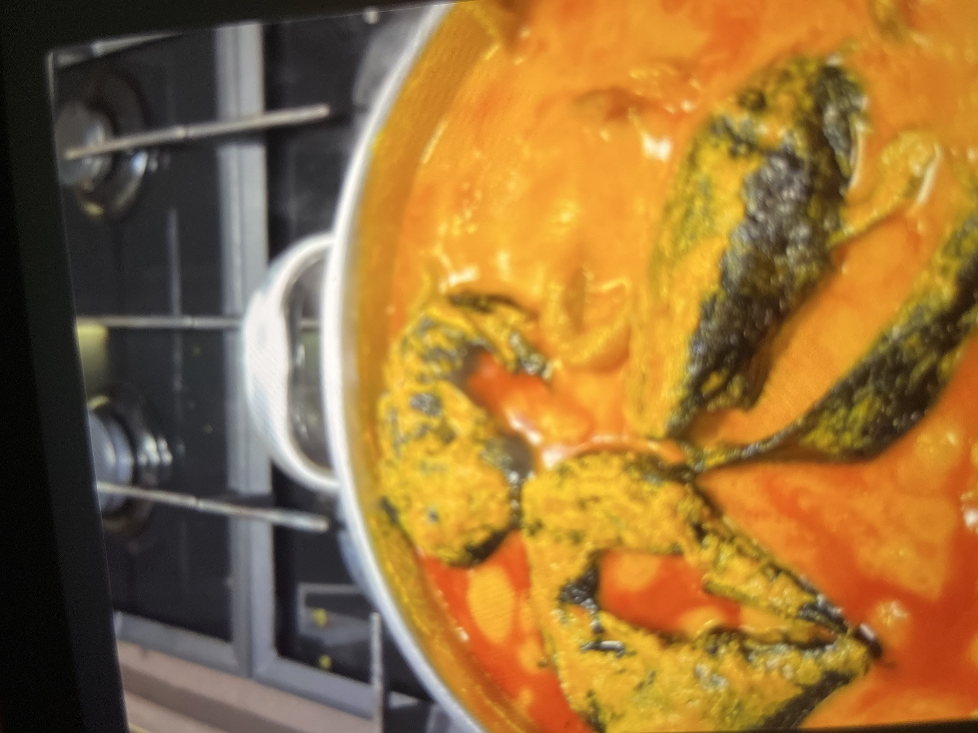 The image shows a close-up, slightly blurry view of a pot of food, likely a stew, with pieces of fish or other seafood submerged in a bright orange-yellow sauce. The food is set against a dark background that appears to be a stovetop with metallic grates, suggesting it is being cooked. The lighting is warm and diffuse. There are no people visible in the image. The scene is consistent with a home kitchen or a restaurant kitchen in Abuja, Nigeria, where such stews are commonly prepared.