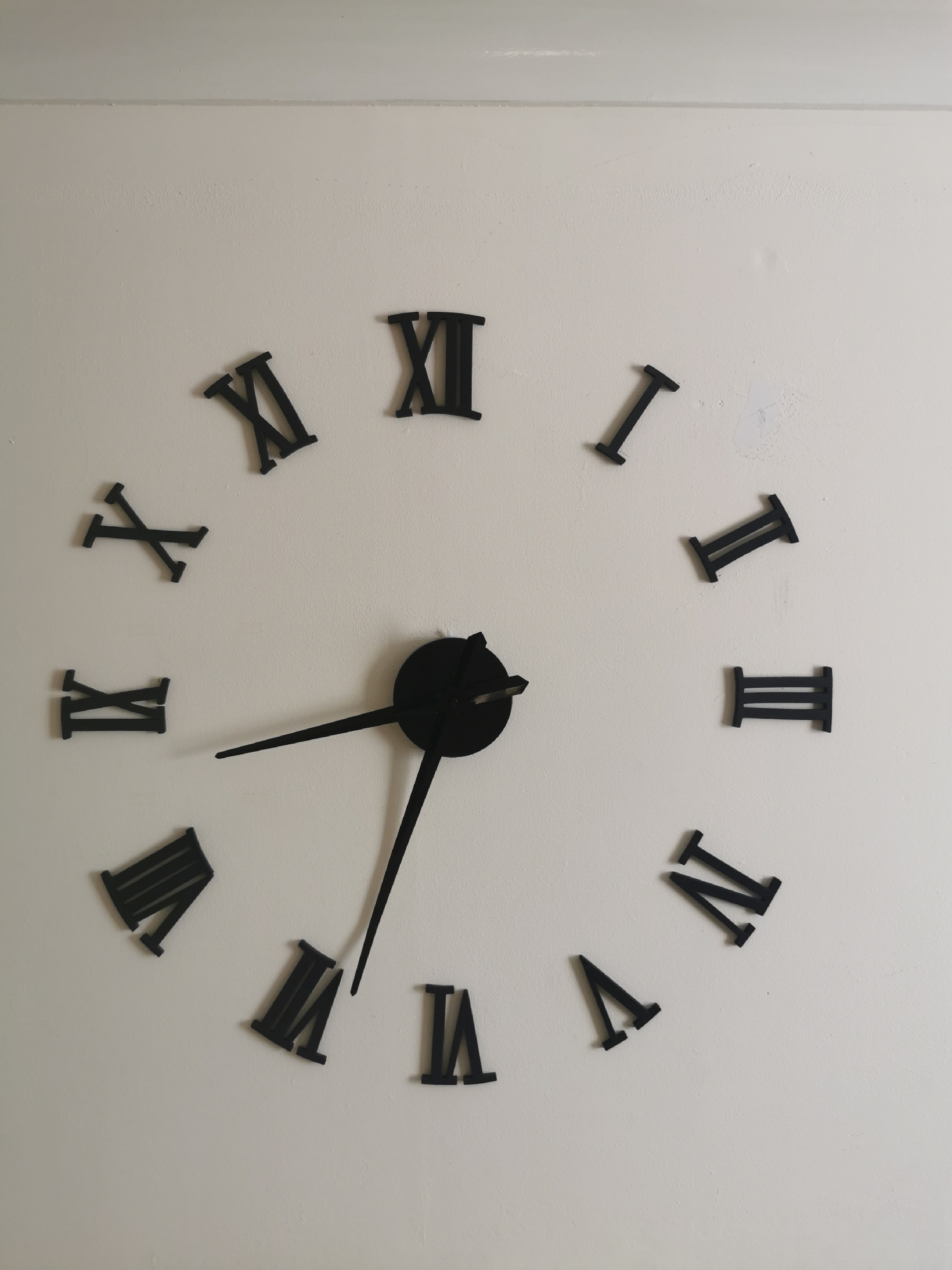 The image shows a close-up, eye-level, interior, medium shot of a black Roman numeral wall clock against a light-colored textured wall. The clock face is not visible, only the individual Roman numeral hour markers and the black clock hands. The Roman numerals are positioned around the clock face, with the hour hand pointing towards the VII (seven) and the minute hand pointing towards the XII (twelve). The clock appears to be a decorative, modern piece. The wall has some minor imperfections, such as a slight discoloration and some textured bumps. The lighting is even, suggesting artificial light or a well-lit room. There are no people or other objects visible in the frame. The location is not specified, but the scene depicts a domestic or commercial interior.