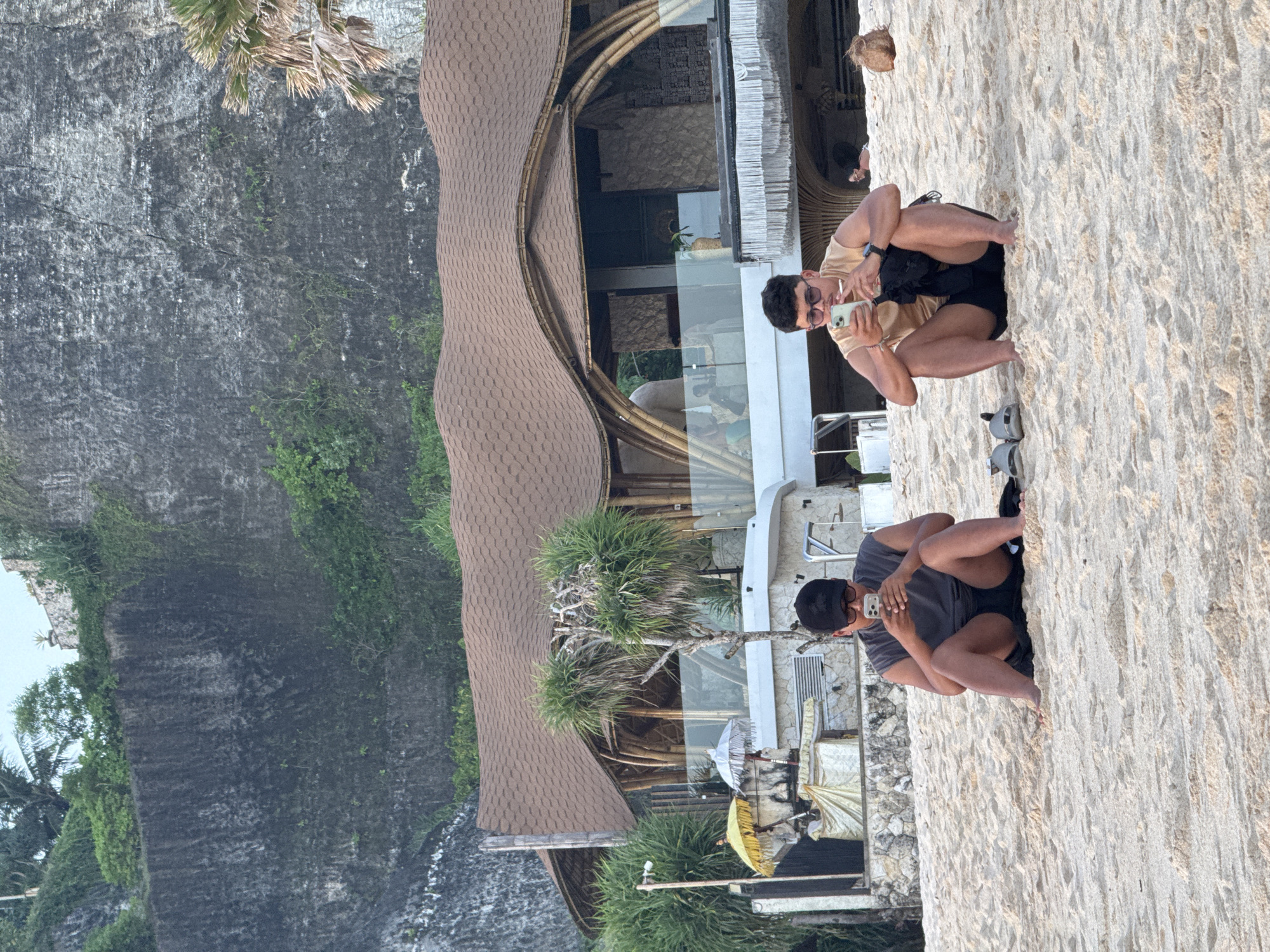 The image captures a scene at a coastal location, possibly a beach, in Kuta Selatan, Indonesia. A large rock cliff partially covered with vegetation is visible to the left. Adjacent to the cliff is a building with a distinctive roof structure, constructed with bamboo and thatched materials. It appears to be a resort or restaurant. On the sandy area in front of the building, two men are seated in similar poses, both looking at and interacting with their phones. One man wears a light brown shirt and dark shorts, while the other wears a dark gray shirt and black shorts. Footwear is placed near the lower man. The presence of beach umbrellas and other resort-style features indicates this is a tourist-oriented setting.