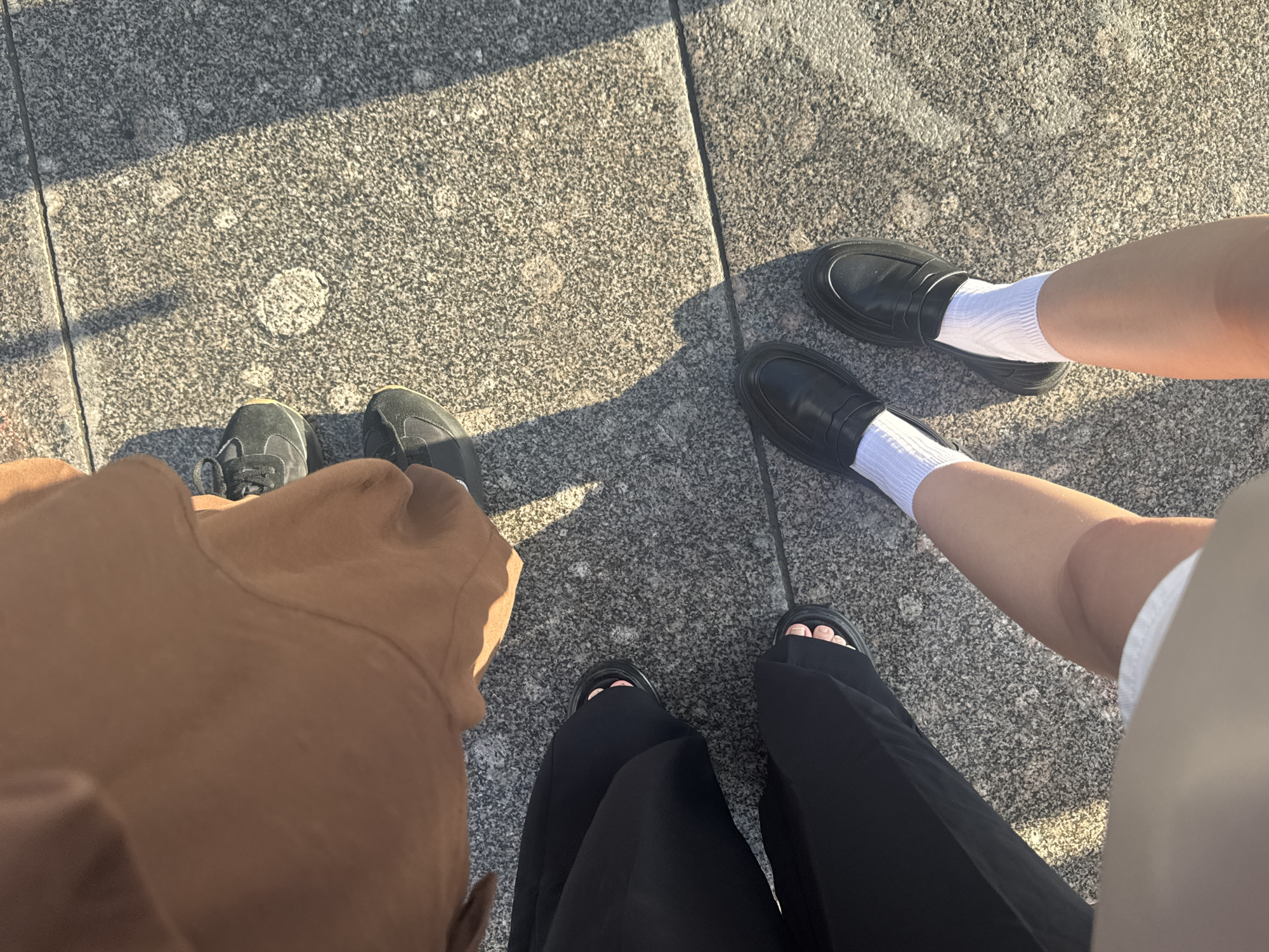The image shows a ground-level view of two individuals' legs and feet. The person on the left is wearing black wide-leg pants and black sandals, revealing their toes. The person on the right is wearing black loafers with white socks and has their legs exposed from the knee down. Both individuals are standing on a speckled gray stone surface, likely a sidewalk or plaza. Sunlight casts shadows on the ground, suggesting it is daytime. The scene does not provide any specific visual cues to pinpoint a location in Saint Petersburg, Russia.