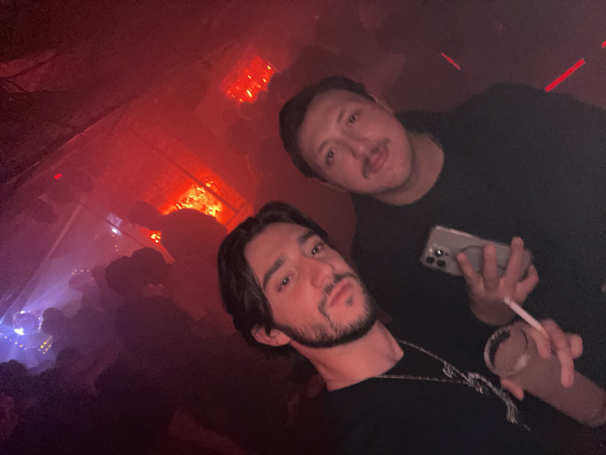 Two men are posing for a selfie in a dimly lit, crowded space with red lighting, potentially a nightclub or bar. The man in the foreground is holding a beverage in one hand and a cigarette in the other, while the man behind him is holding a smartphone. The background includes silhouettes of other people, and a possible lighting fixture is visible near the top left corner of the image. Based on the red lights, the location is likely a nightclub in Mexico City, Mexico.
