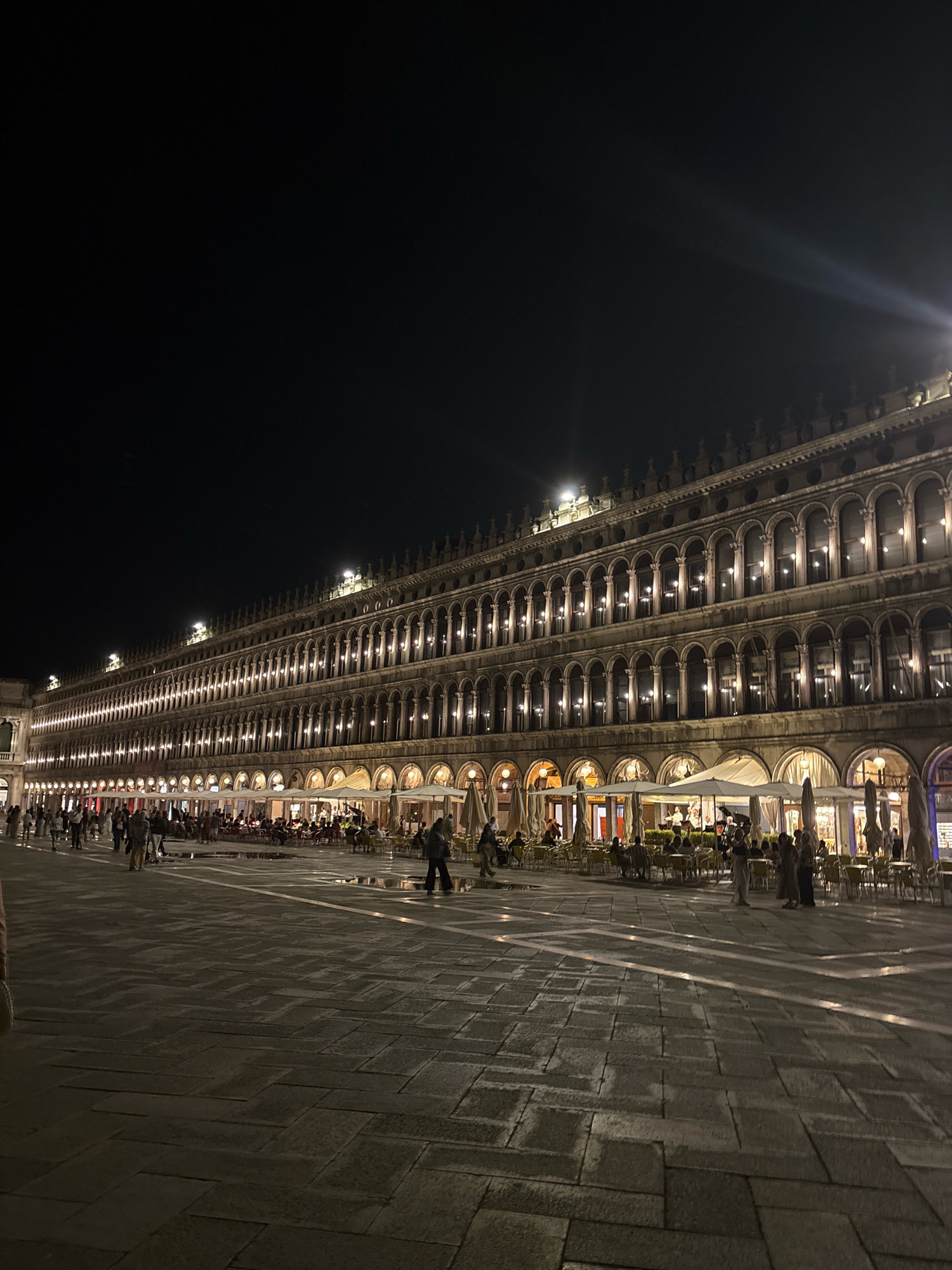 The image captures a nighttime scene in St. Mark's Square (Piazza San Marco) in Venice, Italy. A long, ornate building with multiple levels of arched windows and balconies stretches across the frame, illuminated by warm lights that reflect on the wet cobblestone pavement. The building appears to house cafes or restaurants, as evidenced by the numerous tables and umbrellas set up outside. Several people are visible walking in the square and seated at the outdoor dining areas. The sky is dark, indicating it is nighttime, and a bright light source is visible in the upper right portion of the image, casting a glare. The pavement is wet, suggesting recent rain or that the square has been recently cleaned. The overall atmosphere is one of a lively urban square after dark.
