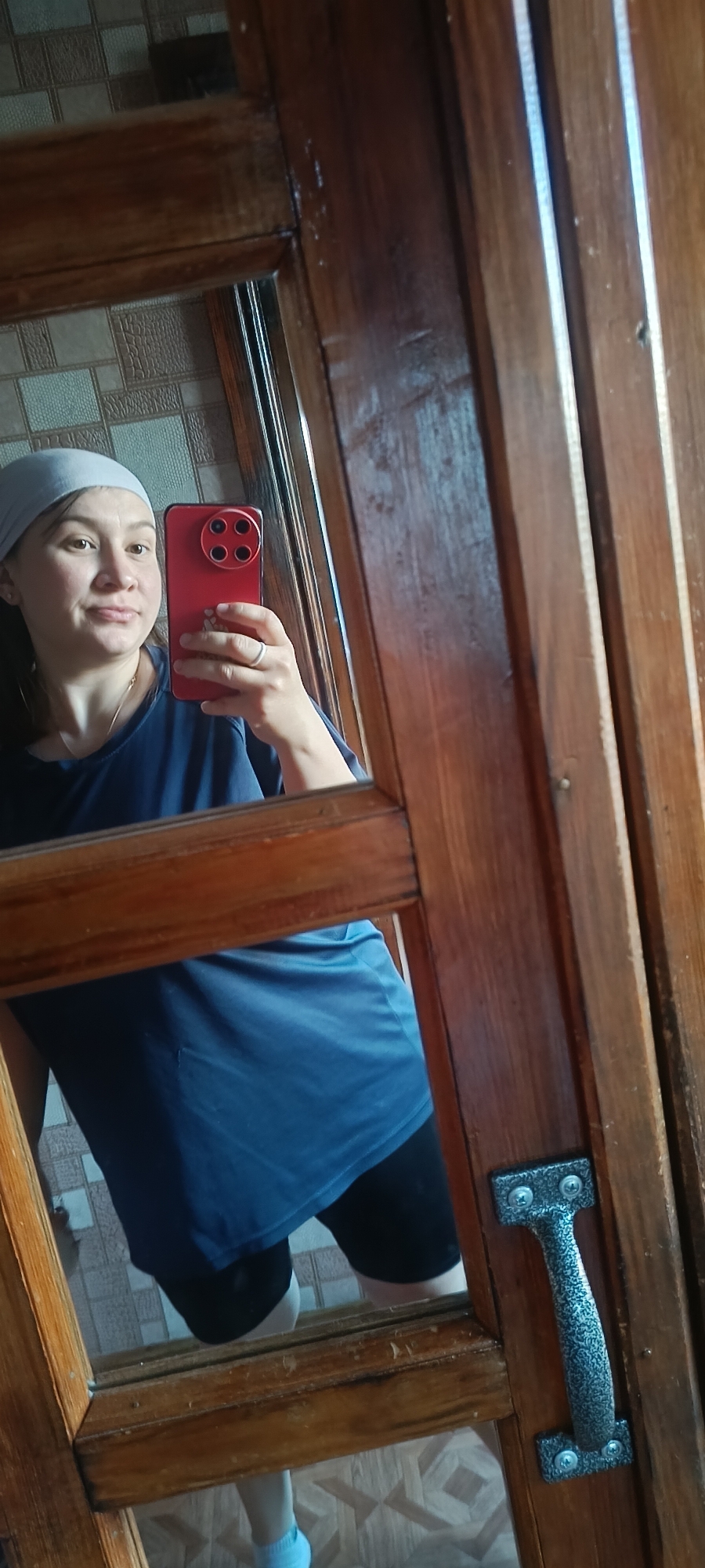The media file is a vertical, medium shot selfie taken in a mirror. The main subject is a woman with fair skin, brown hair, and brown eyes, wearing a light blue bandana and a navy blue t-shirt. She is holding a bright red smartphone in her right hand, which is angled to capture her reflection. The woman is looking at the camera with a slight smile and her head tilted. She is wearing a ring on her left ring finger.
The setting appears to be indoors, likely a home, with a wooden framed mirror. The background reflected in the mirror shows a tiled wall with a geometric pattern in beige and white tones. The woman is wearing black shorts and light blue socks, which are visible at the bottom of the reflection. The right side of the image is dominated by the wooden frame of the mirror, which has a natural wood grain and a handle with a decorative pattern is attached to it. The lighting suggests it is daytime, with natural light coming from an unseen source, creating soft shadows. The overall mood is casual and personal.
There is no visible text in the media file. The location context provided is "Хохлы, Russia."
