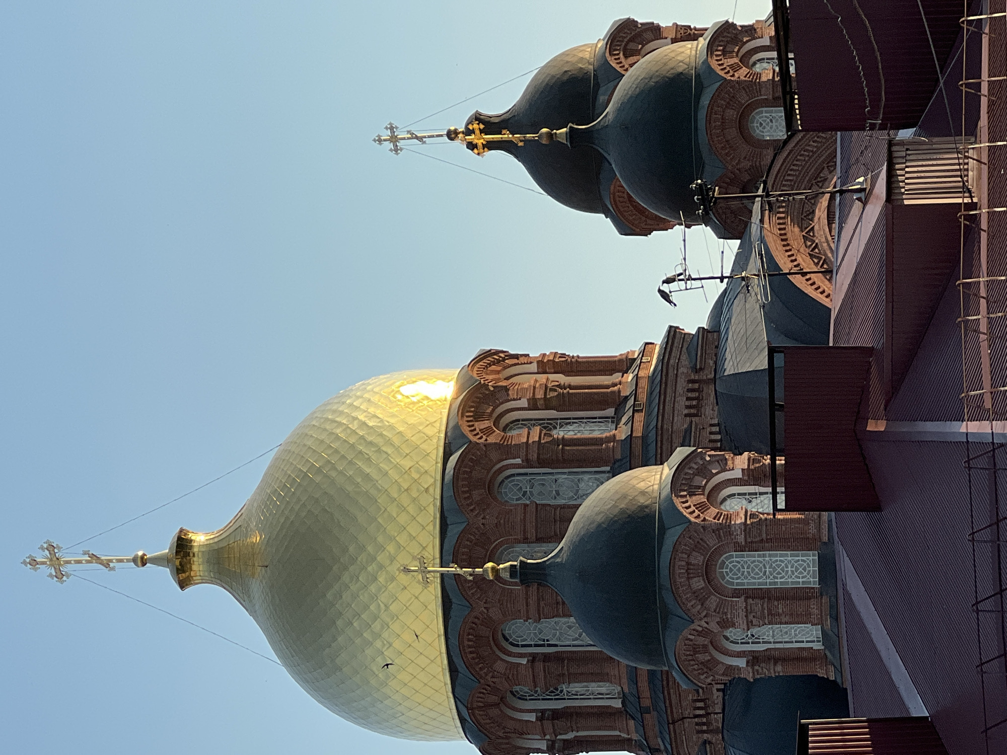This media file shows a close-up, low-angle shot of the exterior of a Russian Orthodox church, with a bright blue, cloudless sky as the backdrop. The main subject is the church's architecture, featuring a large, golden dome with a cross at its apex. Adjacent to the golden dome are several darker, smaller domes, also topped with crosses. The building's facade is constructed from red brick and features intricate detailing, including arched windows with decorative tracery and layered architectural elements. A metallic roof, likely made of corrugated metal, covers a portion of the building. The lighting suggests it is daytime, with the sun glinting off the golden dome, indicating a clear day. Two birds are visible in flight against the sky. The image's orientation is rotated, making the structures appear to be upside down. The setting is identifiable as Krasnodar, Russia. No people are visible in the frame.