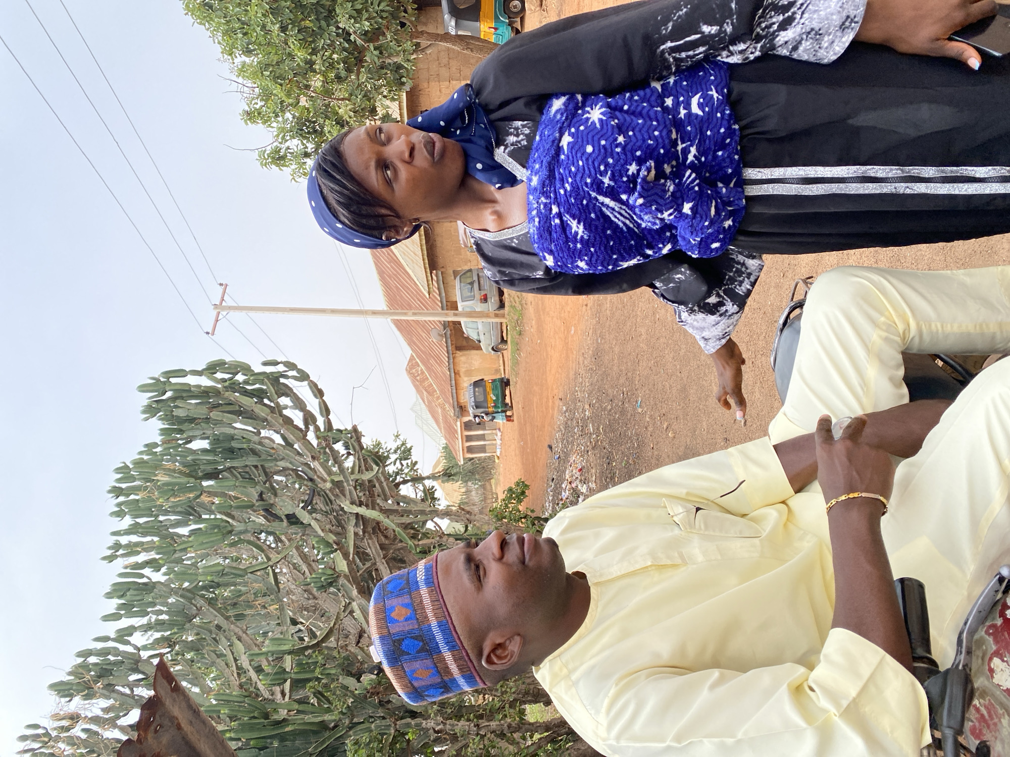 In Kuru Karama, Nigeria, a man wearing a light yellow shirt and a blue and brown kufi sits on a vehicle, possibly a motorcycle. He is wearing a gold bracelet and appears to be in conversation with a woman standing beside him. The woman is wearing a black and white patterned garment and has a blue headscarf. She has a blue wrap secured around her torso. The background shows a dirt road with a building and a motorized tricycle. There are power lines and a large cactus-like plant on the left side of the image. The sky is overcast.