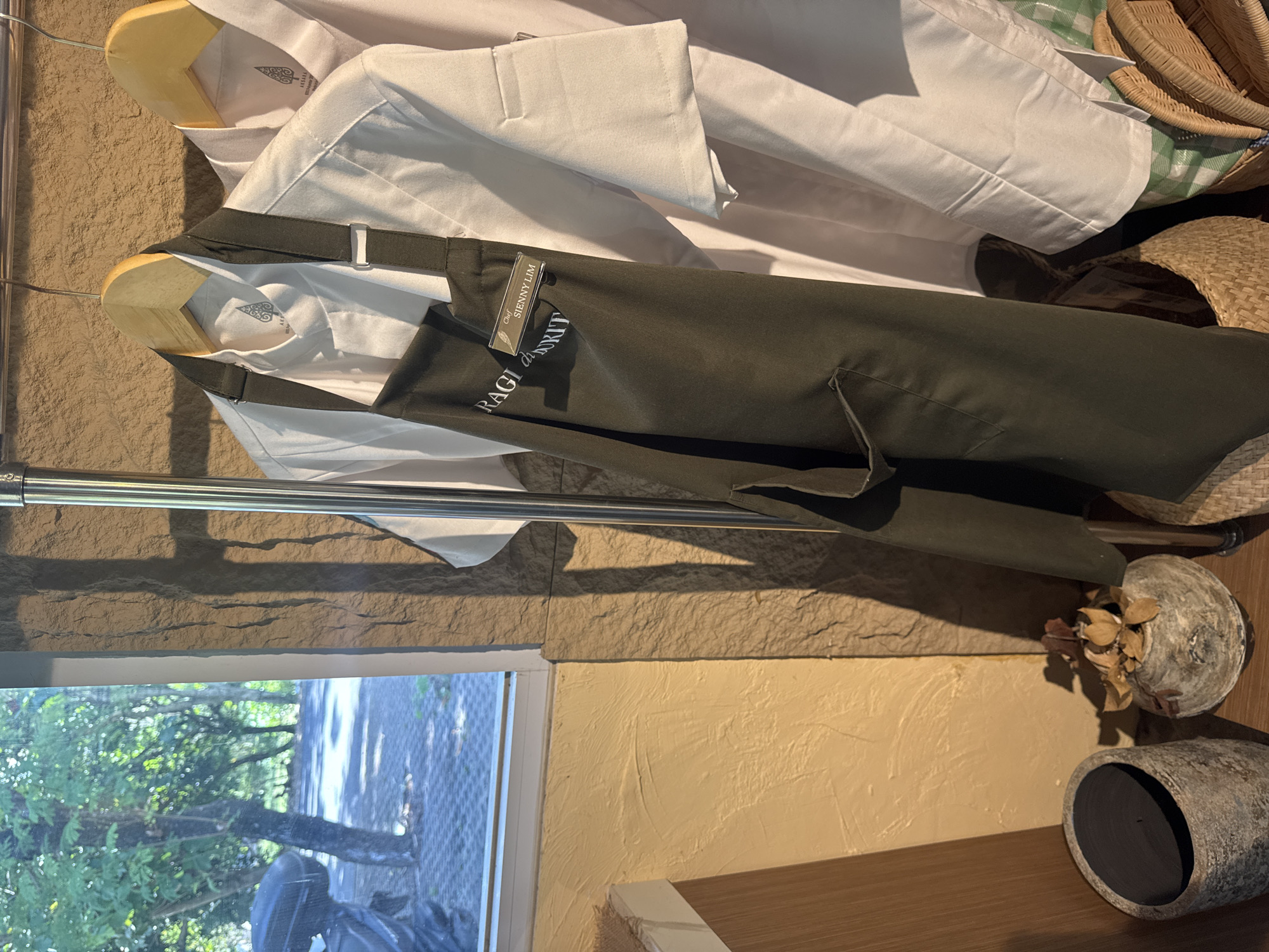 The image presents a chef's attire hanging in what appears to be an indoor space, likely in Kuta Selatan, Indonesia. Visible on a clothing rack are at least two white chef coats, and a dark green or olive apron. The apron bears a name tag that reads "Chef SIENNY LIM" over the word "RAGI". The garments are hung on wooden hangers. The background includes a light brown textured wall, a window with green foliage visible outside, and a shelf with a decorative planter. Additionally, various woven baskets are present to the right of the clothing rack. The scene is illuminated with indoor lighting.