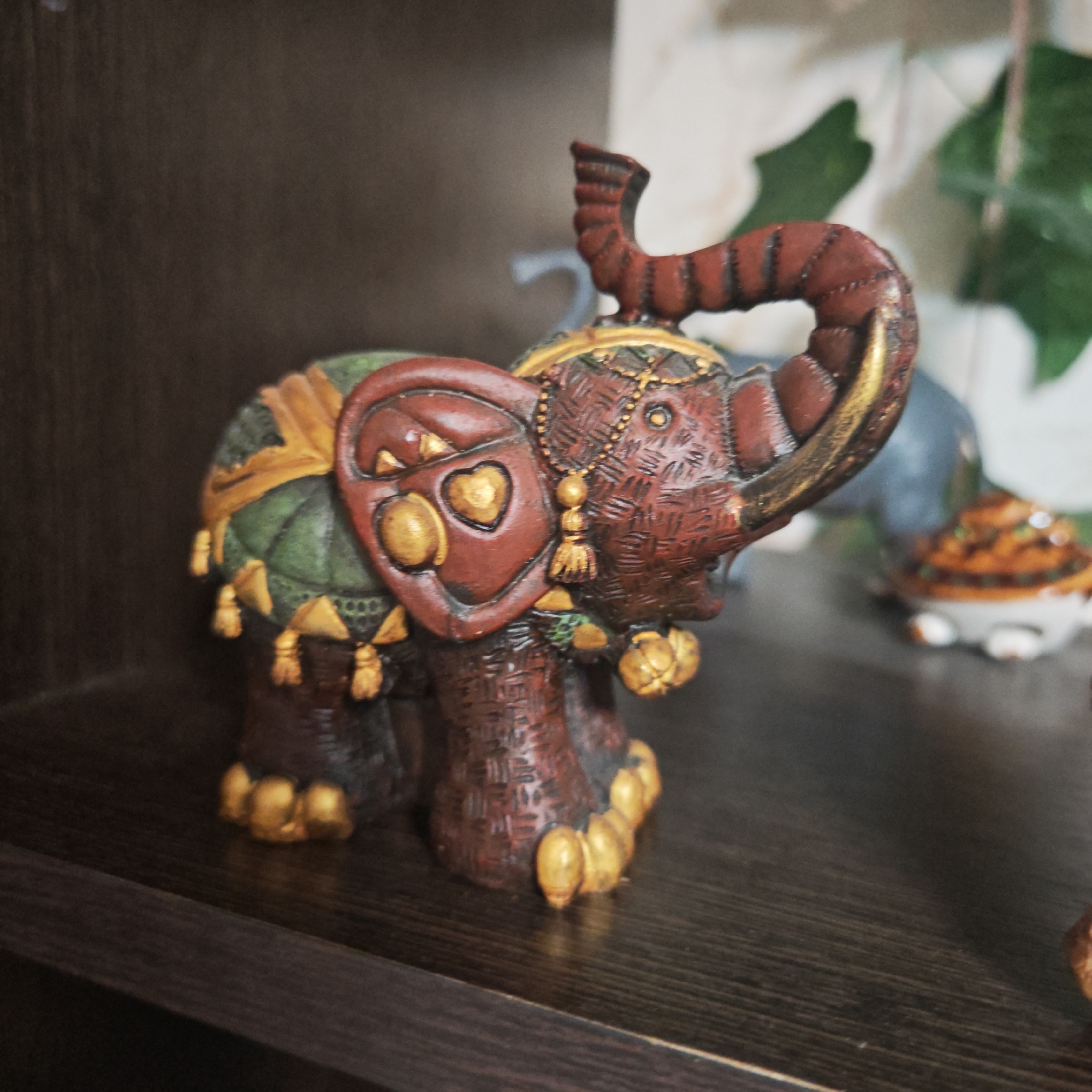 The image features a decorative elephant figurine positioned on a dark brown wooden surface. The figurine is primarily brown with gold accents on its feet, trunk, ears, and decorative elements on its back. A green cloth is draped on its back, with gold tassels hanging from it. The elephant's ears have heart-shaped decorations with gold accents. The environment includes hints of other decorative items and a plant in the background, but they are out of focus. It is not possible to confirm the location to Rostov, Russia from this image.