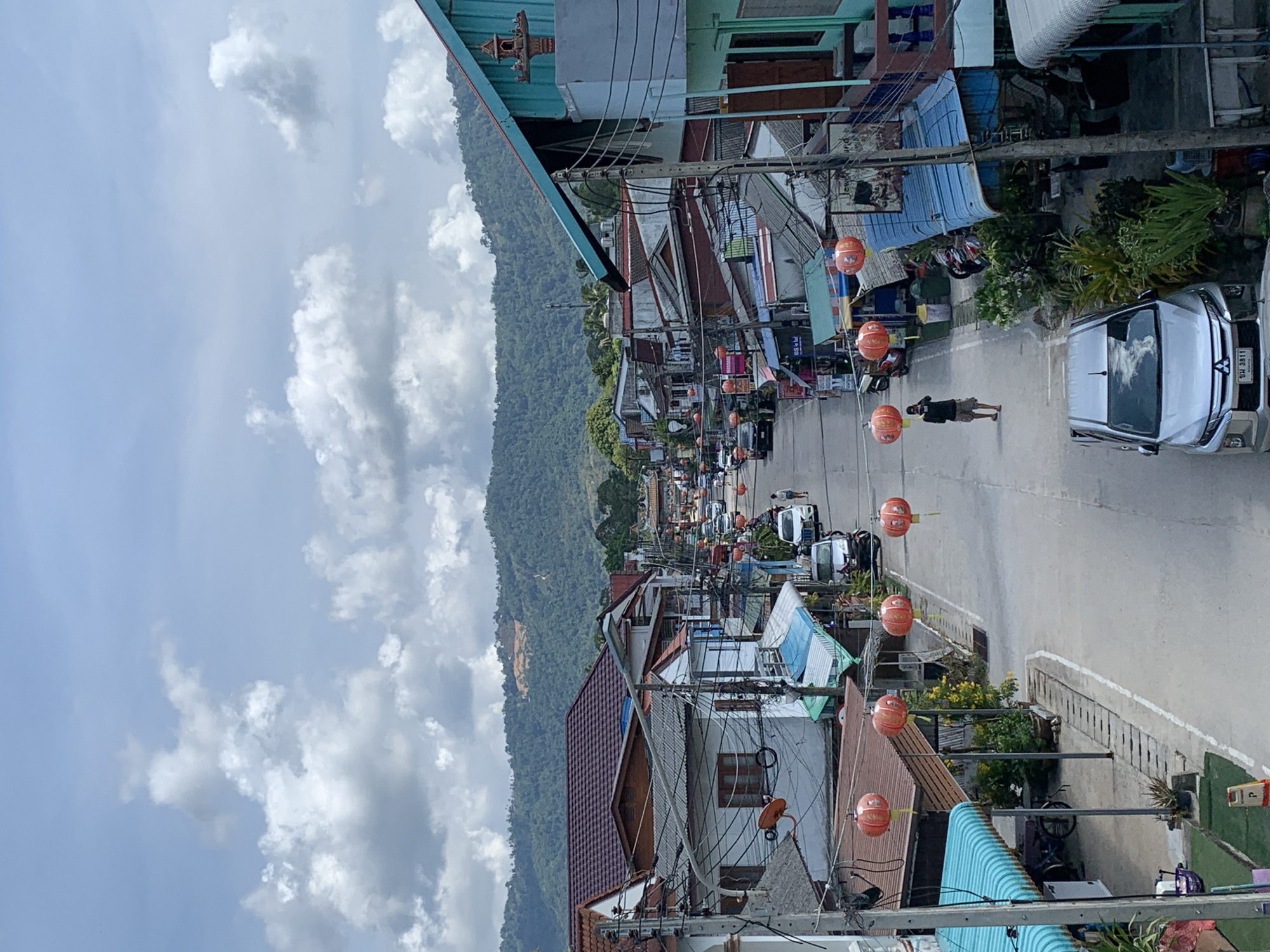 The image depicts a street scene in Baan Mae Nam, Thailand. The street is lined with buildings of varying heights and colors, with power lines crisscrossing overhead. Lanterns are strung across the street. A person is walking on the right side of the road, near a parked silver car. On the left, a mountainside rises. The sky is cloudy.