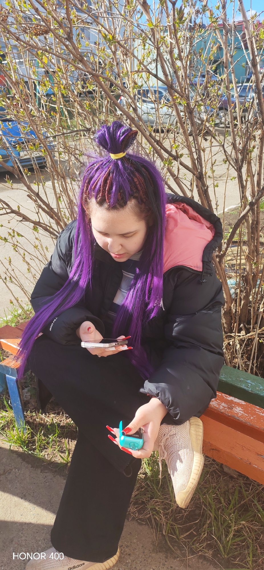 A young woman with purple braided hair and long red nails sits on a bench outdoors, likely in a park or public area. She is wearing a black puffer jacket with a pink hood, black pants, and beige Adidas shoes. She is holding a cell phone in one hand and a small, turquoise object (possibly a vape or small electronic device) in the other. The background shows a leafless bush or small tree with some budding leaves. In the distance, there is a glimpse of a building and vehicles, suggesting an urban environment. The text "HONOR 400" is visible in the bottom left corner of the image. The location may be in the city of Noviy, Russia.