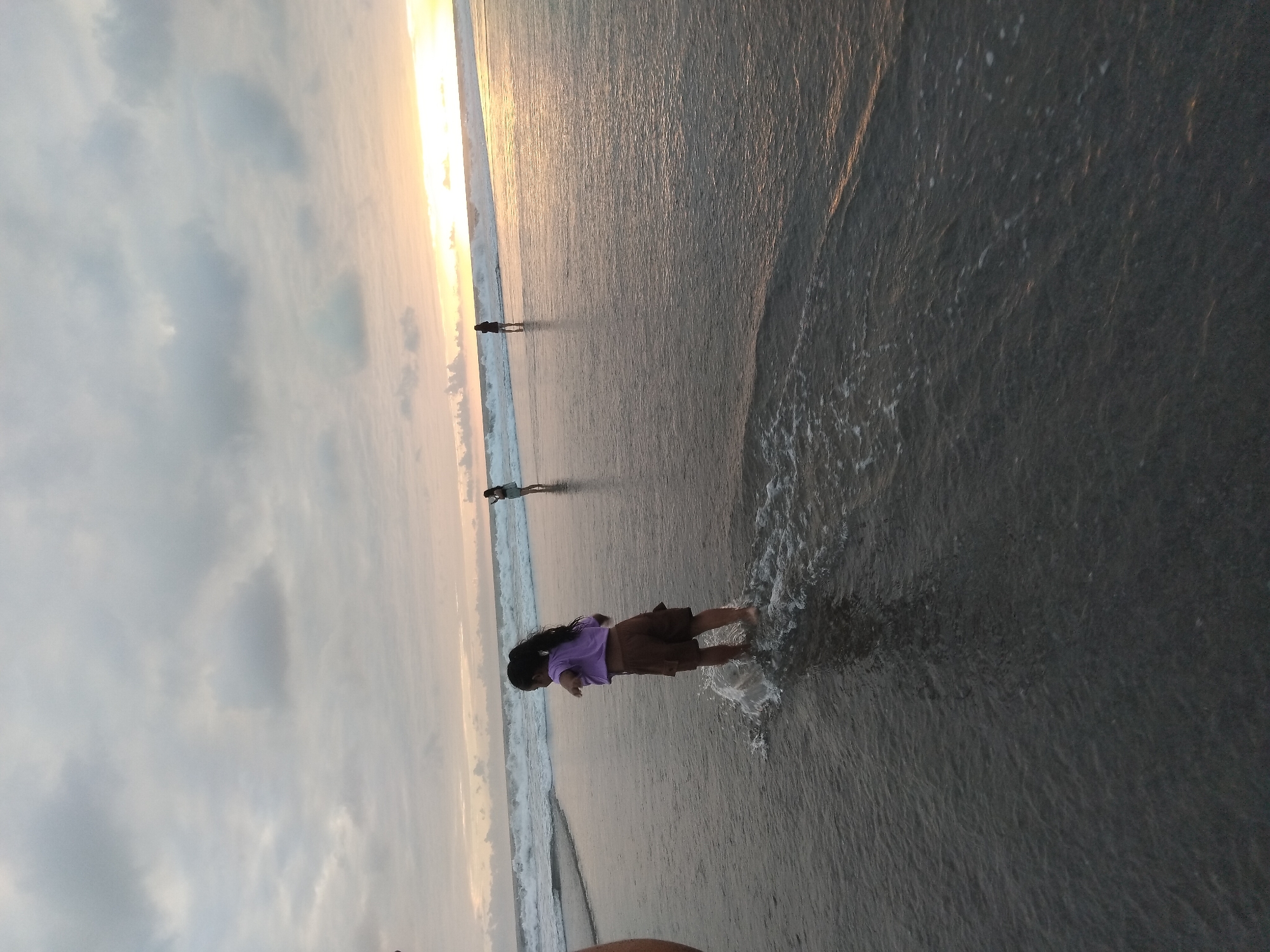 A young girl with long dark hair and wearing a purple top and brown shorts stands in the shallow water at the edge of a beach, splashing. In the distance, two other figures are visible standing in the water, and the ocean waves are breaking. The sky is overcast with clouds, but the setting sun casts a warm glow on the horizon. The scene appears to be located in North Kuta, Indonesia, a popular tourist destination known for its beaches. The overall atmosphere is serene and peaceful, evoking a sense of summer vacation and family fun.