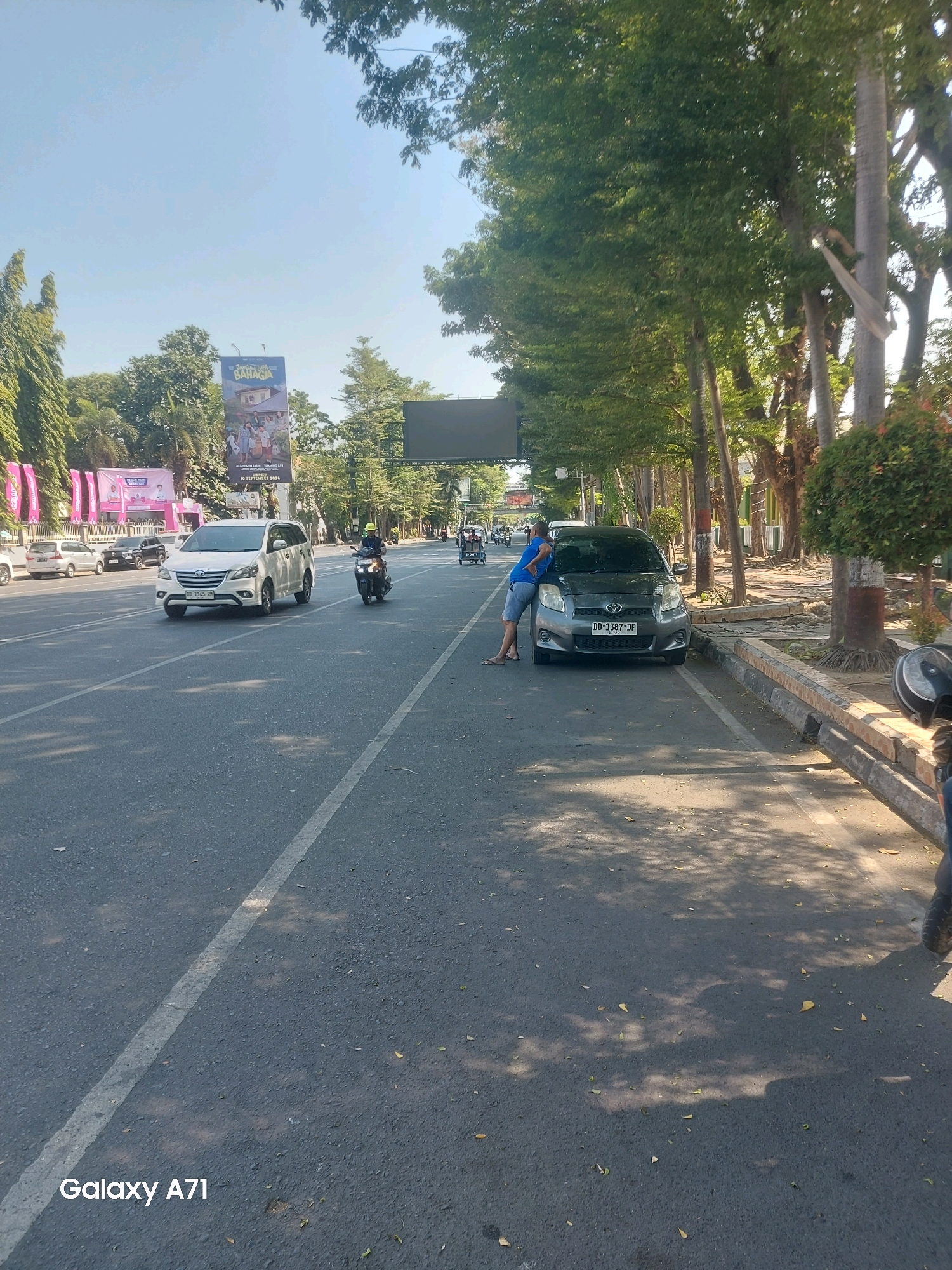 A daytime, outdoor, eye-level shot captures a street scene in Makassar, Indonesia. The foreground is dominated by an asphalt road with a white lane marker. To the right, a person in blue shorts and a blue t-shirt leans against a grey hatchback car, parked on the side of the road. The car has a license plate reading "DD 1387 DF". In the background, trees line the street, casting dappled shadows on the road. Further back, several vehicles are visible, including a white minivan and a motorcycle with a rider wearing a yellow helmet. A billboard advertising "BAHAGIA" is also present. The sky is clear and blue.