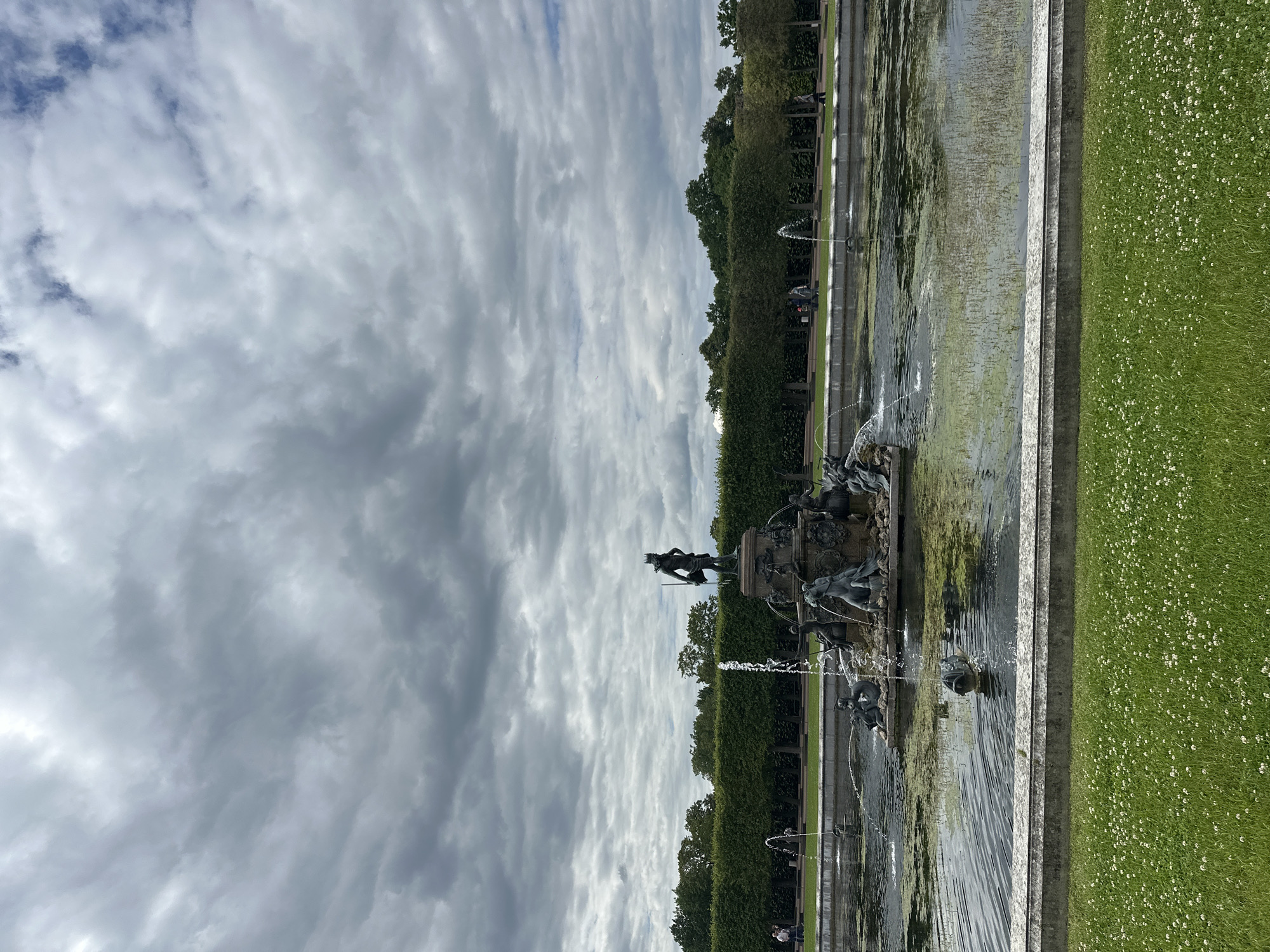 The image shows a fountain in Peterhof, Russia. The fountain is a large, multi-tiered sculpture with several figures, including a central figure of a man with a crown and a trident. Water is spraying from the fountain and into the surrounding pond. The pond is surrounded by a manicured hedge and a grassy lawn with small white flowers. In the background, there are trees and a cloudy sky. Several people are visible walking in the distance. The fountain appears to be part of a larger park or garden complex.