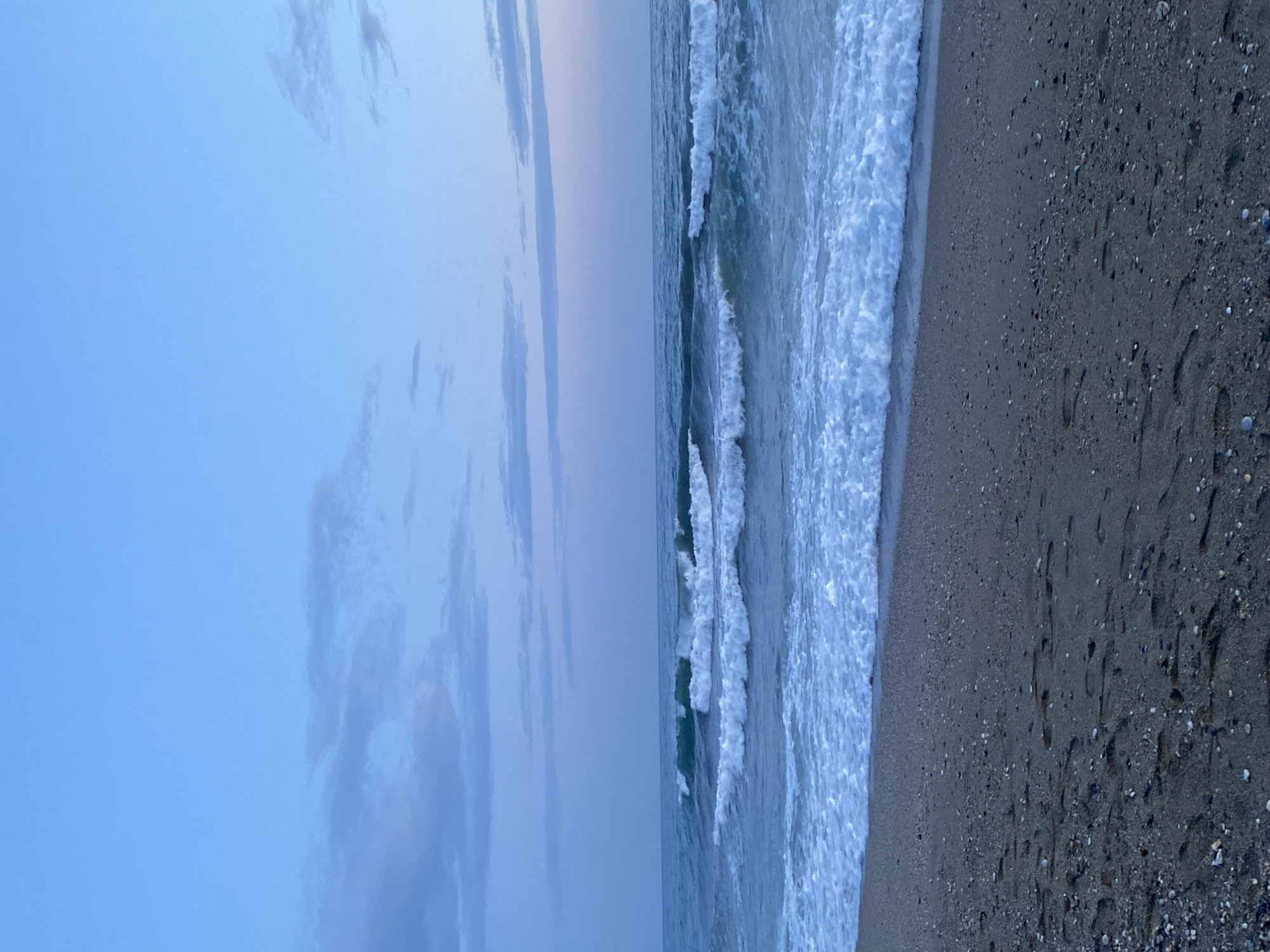The image depicts a beach scene at Alanya, Turkey. The sky is a gradient of light blue to a hazy pale purple near the horizon, with sparse, elongated clouds. The sea has gentle waves breaking onto the shore, creating white foam. The sand is dark and damp, with visible footprints and small pebbles. The overall scene is tranquil and captures the transition between day and night.