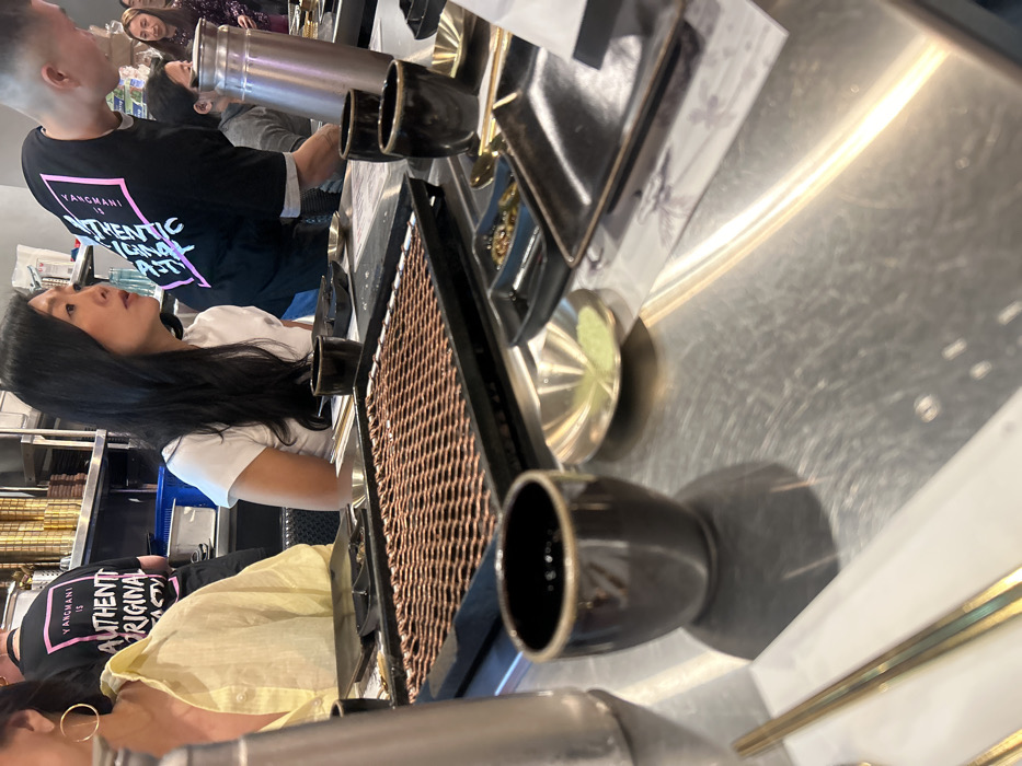 A scene at a restaurant features a long metal table with a brown grill and various dark cups arranged along its edge. Two women wearing light-colored shirts, and a man in a black shirt with pink accents are positioned around the table. In the background, additional people and shelving stocked with items are visible. The image is taken from a high angle and appears slightly tilted.