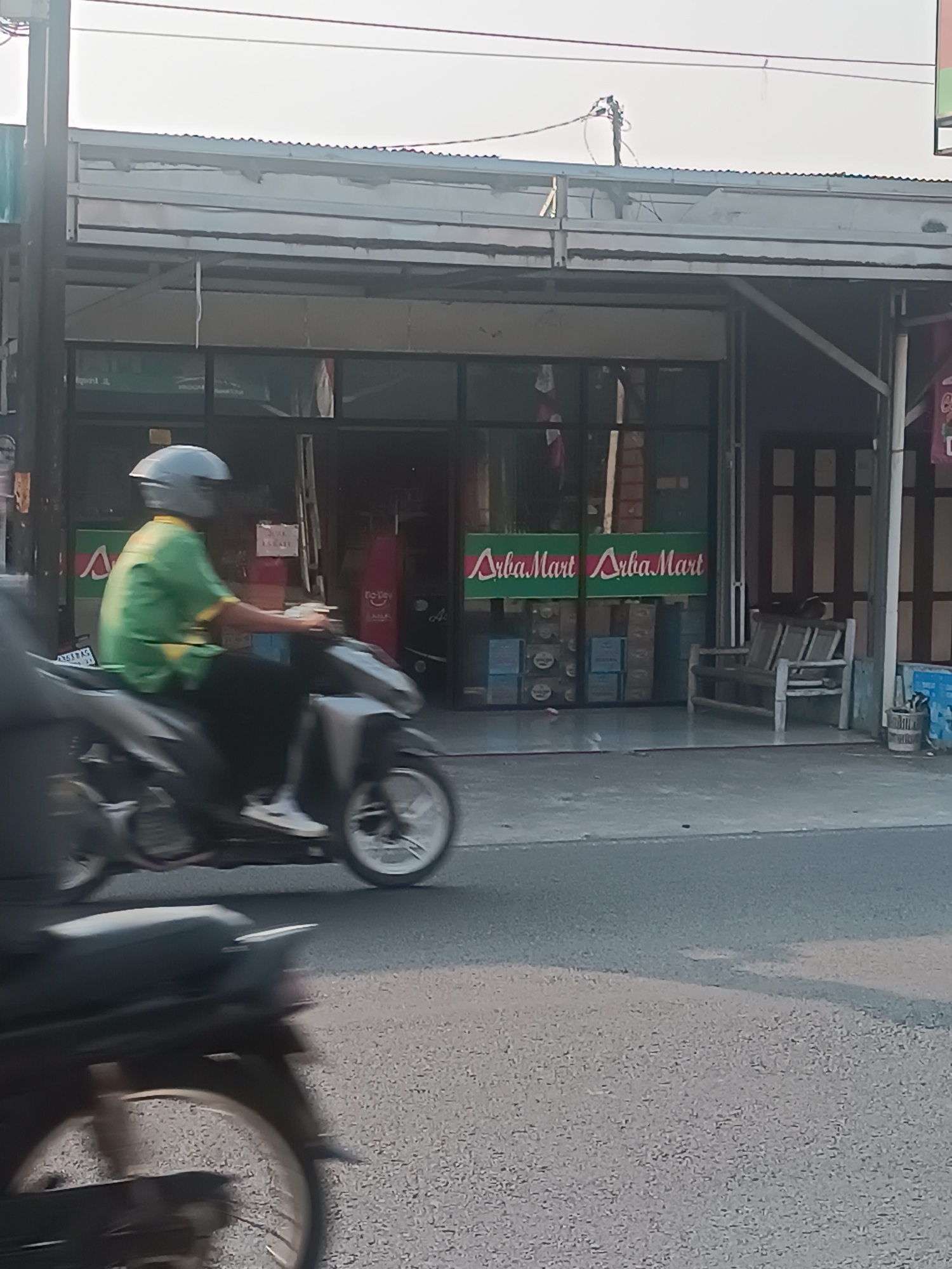 This is a medium-shot, eye-level photograph taken outdoors on a street in Imogiri, Indonesia, likely during daylight hours given the lighting. In the foreground, a person wearing a green shirt and a grey helmet is riding a silver and black scooter from left to right. The scooter is in motion, indicated by the blur of its wheels and the overall motion blur in the image. The rider's legs are visible, and they are wearing white sneakers.
Behind the scooter, a storefront is visible. The store has a glass front and a corrugated metal awning. Two green signs with white text that reads "Arba Mart" are prominently displayed on the storefront. The interior of the store appears to be stocked with boxes and other merchandise. To the right of the storefront, a wooden bench with three seats is placed outside. Further to the right, there are some blue boxes and a cylindrical trash receptacle. The road surface is asphalt with some light gravel. The background shows a building with a striped facade and overhead electrical wires.