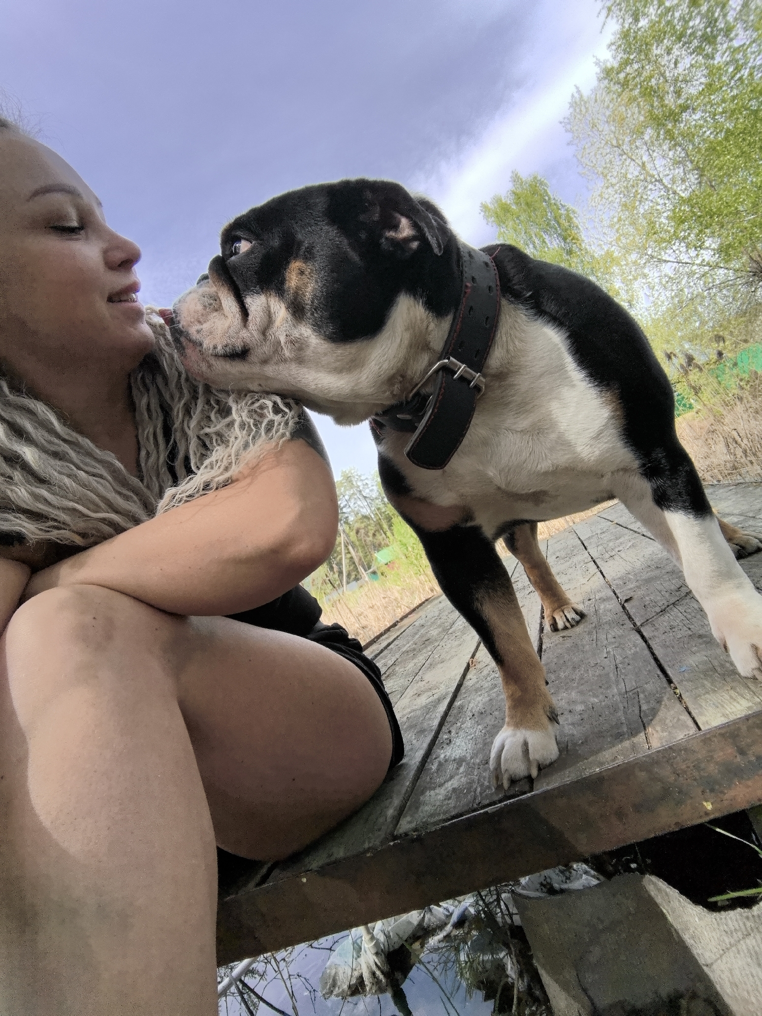 A woman with light-colored dreadlocks is sitting on a wooden deck and is being licked on the face by a black, white, and brown Bulldog. The dog is standing on the deck. The scene takes place outdoors near a body of water. Some trees and foliage are visible in the background. There is some water under the dock. The location is Noviy, Russia.
