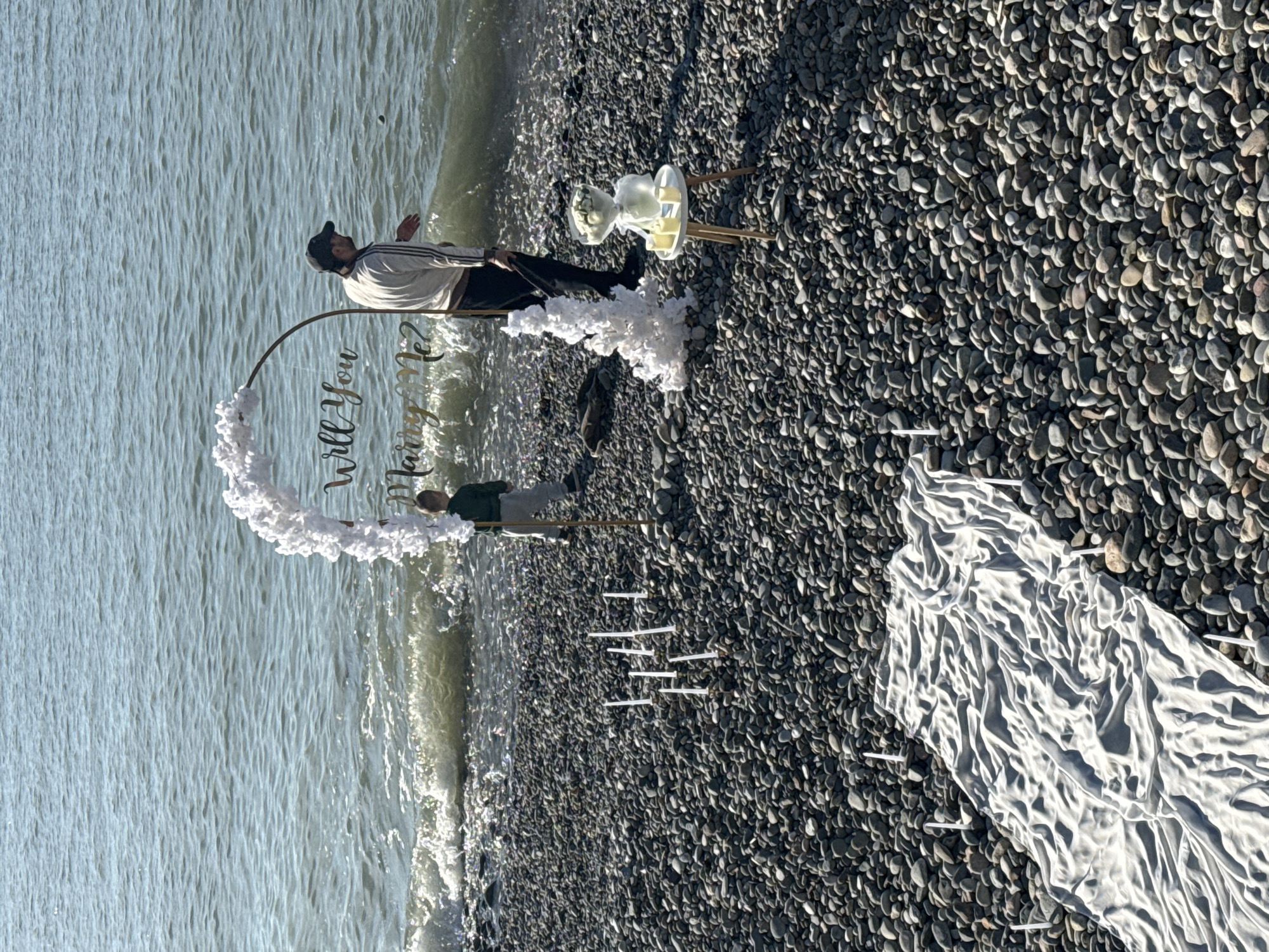 On a pebble beach in Batumi, Georgia, a man wearing a black baseball cap, white and black striped shirt, and dark pants stands near a heart-shaped sign reading "Will You Marry Me?". The sign is decorated with white floral accents. Near the man, a small, white, three-legged table holds a bouquet of white flowers wrapped in plastic. In the foreground, a white fabric runner is lined with white objects, possibly candles. Waves are visible behind the man and the sign. Another person is standing behind the sign.