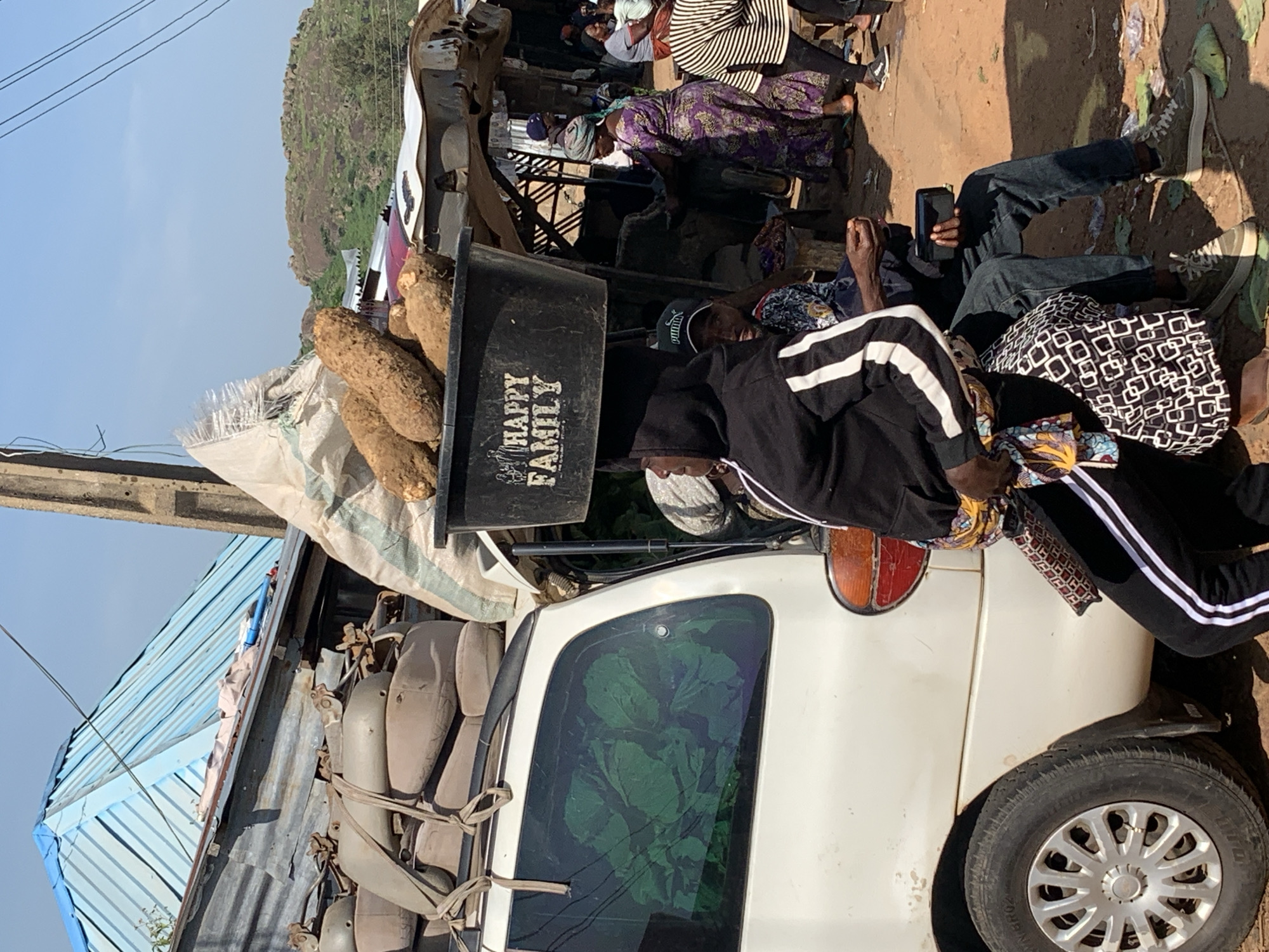 In the city of Jos, Nigeria, a white van is parked in an outdoor market. The van is loaded with goods, including what appear to be large yams and other produce. A black plastic container, labeled "HAPPY FAMILY," sits on top of the van, also filled with yams. Several people are present in the scene. A woman wearing a black hoodie with white stripes is seated, with a colorful fabric wrapped around her waist. Beside her, a man in a blue patterned shirt and a black baseball cap is also seated, holding a dark object that appears to be a smartphone. Other individuals are seen standing and interacting in the background, some dressed in traditional Nigerian attire. The environment suggests a busy marketplace, with various stalls and structures visible. The ground is dirt, and the overall atmosphere is that of daily commerce. The sky is clear and blue, indicating a sunny day.