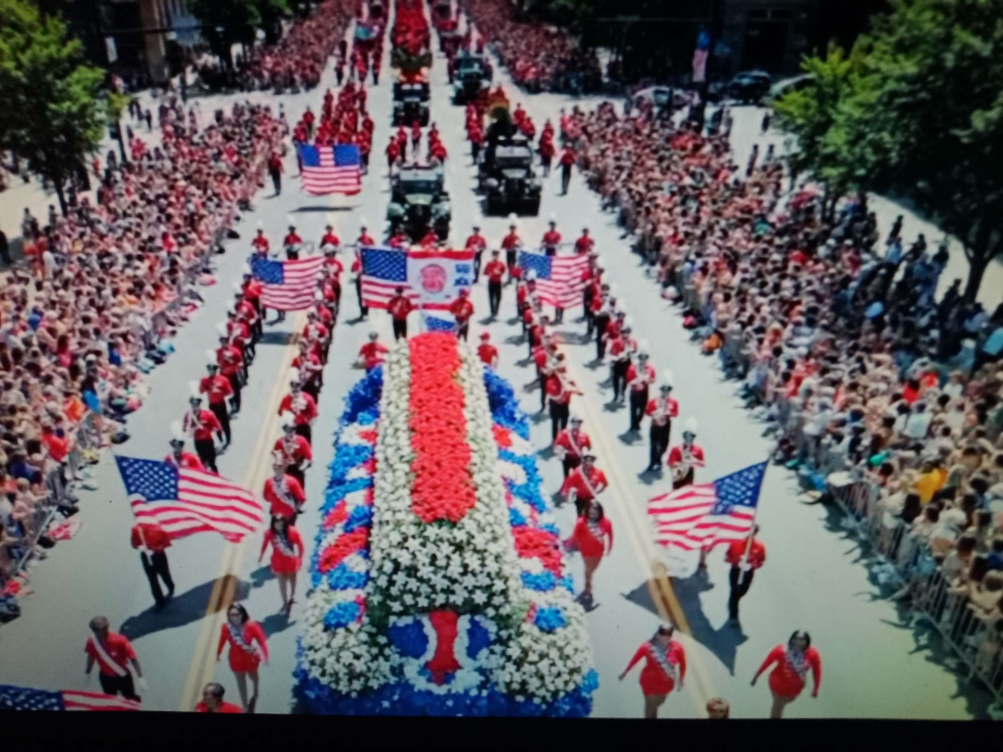 The image depicts a vibrant parade winding through a city street lined with a large crowd of spectators. Several marching bands, identifiable by their red uniforms and white hats, are prominently featured, carrying large American flags. The parade also includes a large, elaborately decorated float covered in red, white, and blue flowers, possibly representing a patriotic theme. Several vehicles, possibly military vehicles, are also part of the procession. The spectators, dressed in casual summer attire, are densely packed along both sides of the street, suggesting a significant public event. Trees and buildings are visible in the background, indicating an urban setting. There is no information in the image or provided context to suggest the location is in Chiajna, Romania.