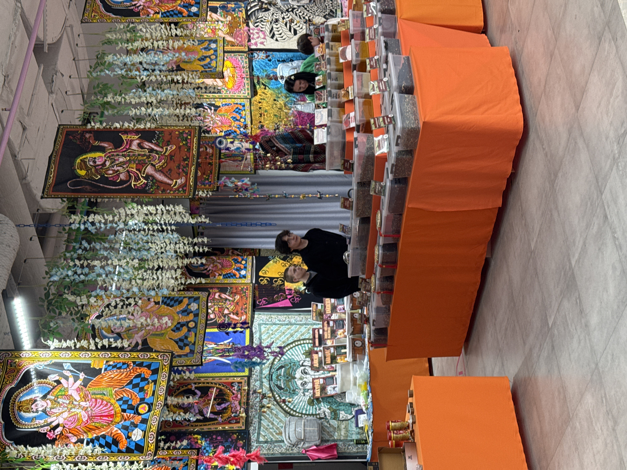 This image shows an indoor marketplace in Perm, Russia. Two people are standing at a stall covered in orange fabric, which features numerous bins of goods. The backdrop of the stall is decorated with colorful tapestries depicting deities and floral garlands. The floor is tiled.