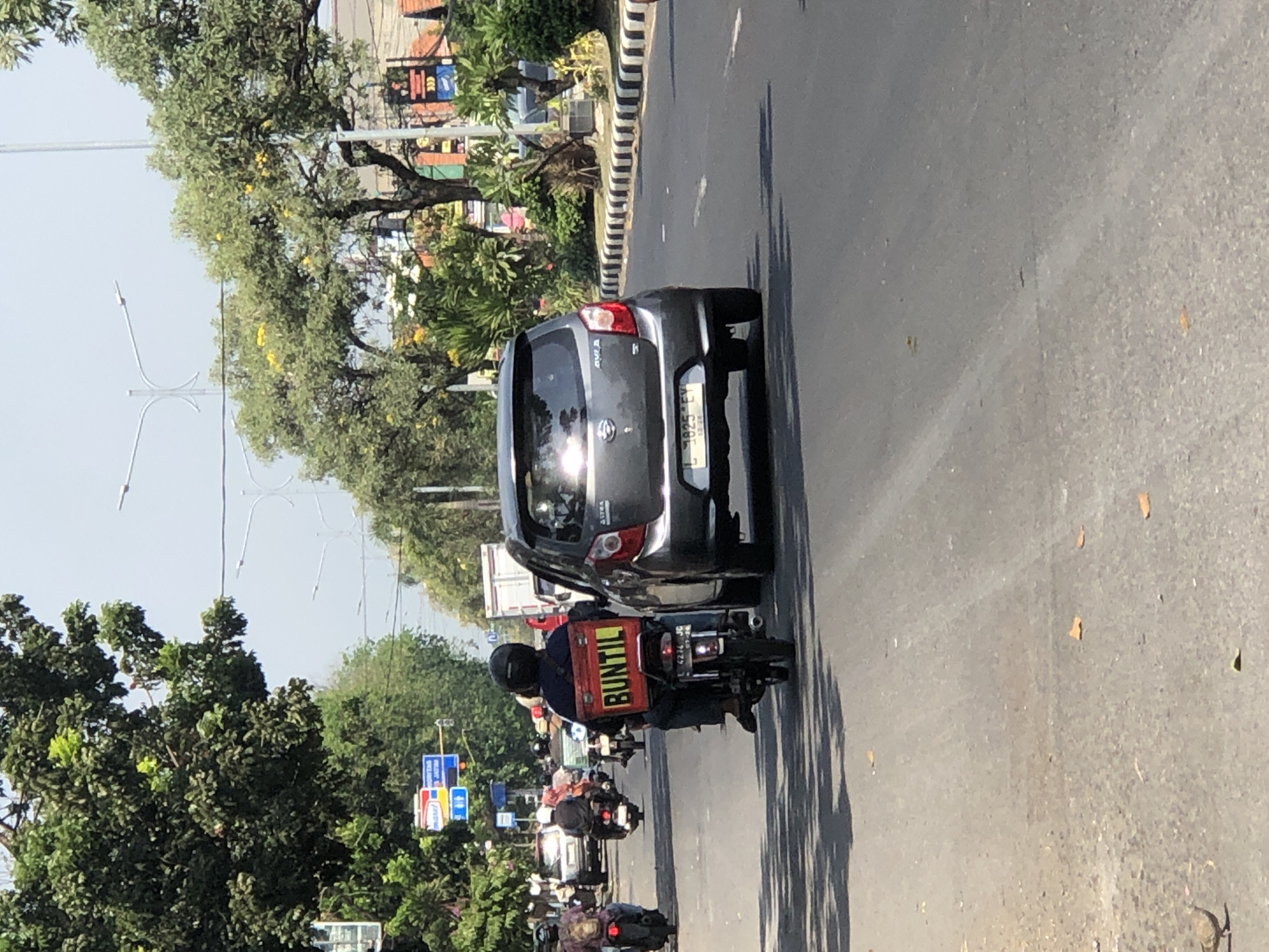 The image captures a street scene in Larangan, Indonesia, under a bright, clear sky. In the foreground, a motorcycle rider wearing a black helmet and dark clothing is seen from behind. Attached to the rear of the motorcycle is a bright red box with "BUNTIL" printed in yellow capital letters. The motorcycle's license plate reads "L 4274 JG". The rider is positioned on the right side of a two-lane road.
To the right of the motorcycle, a dark gray hatchback car is visible, facing away from the viewer. Its license plate appears to be "L 1825 EY".
Several other motorcycles and a few cars are present on the road, suggesting moderate traffic. Trees with lush green foliage line the sides of the road, with some trees bearing yellow flowers. Streetlights and traffic signals are also visible in the background. The road surface is asphalt, with painted lane markings and a striped curb on the right. Fallen leaves are scattered on the road. The overall impression is a typical urban street scene during daylight hours.